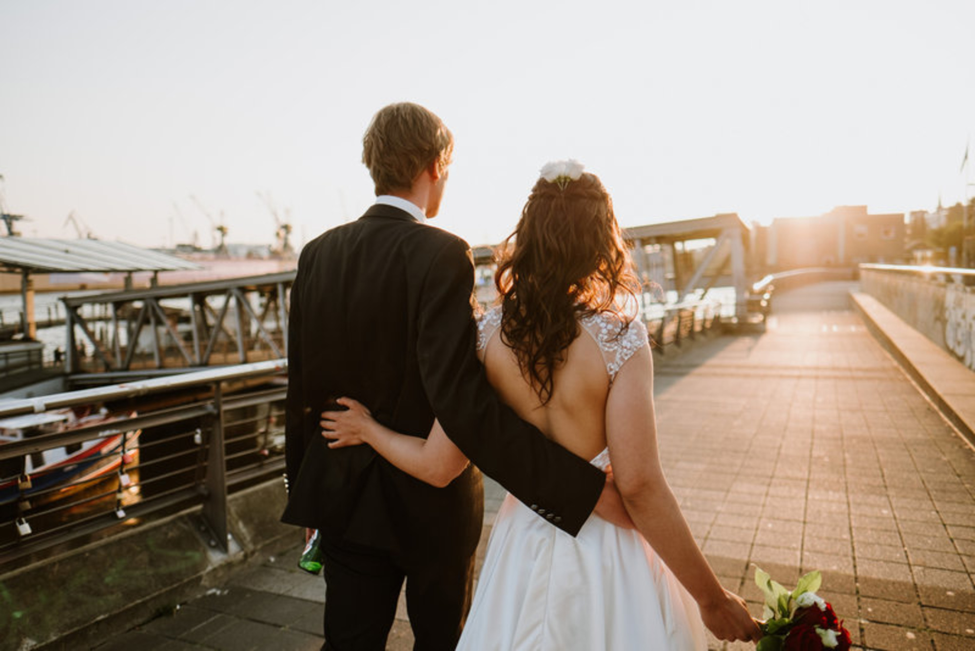Newlywed couple walks arm in arm along a waterfront promenade at sunset holding a bouquet.