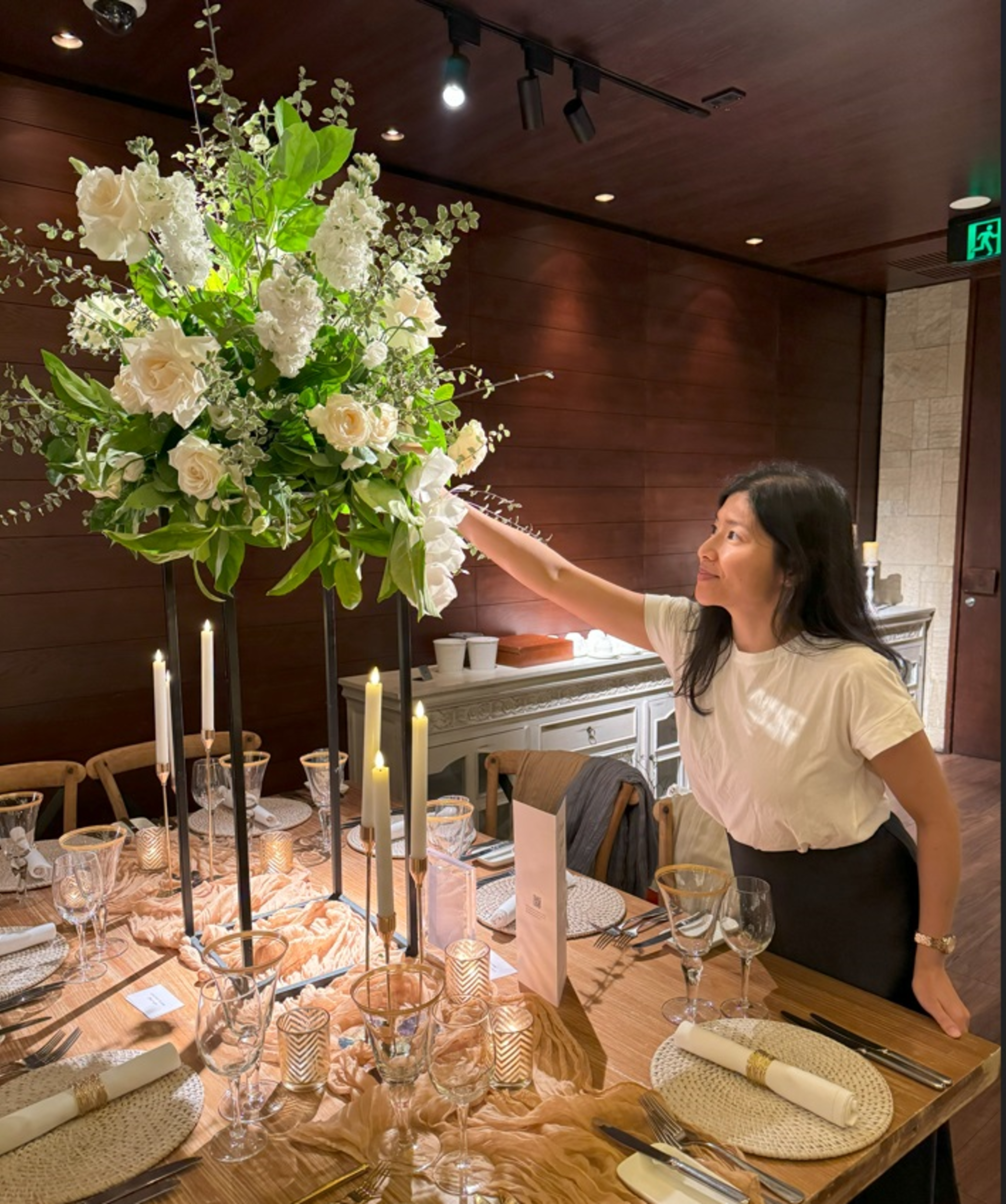 Florist adjusts a tall white and green floral centerpiece on an elegant candlelit wedding reception table.