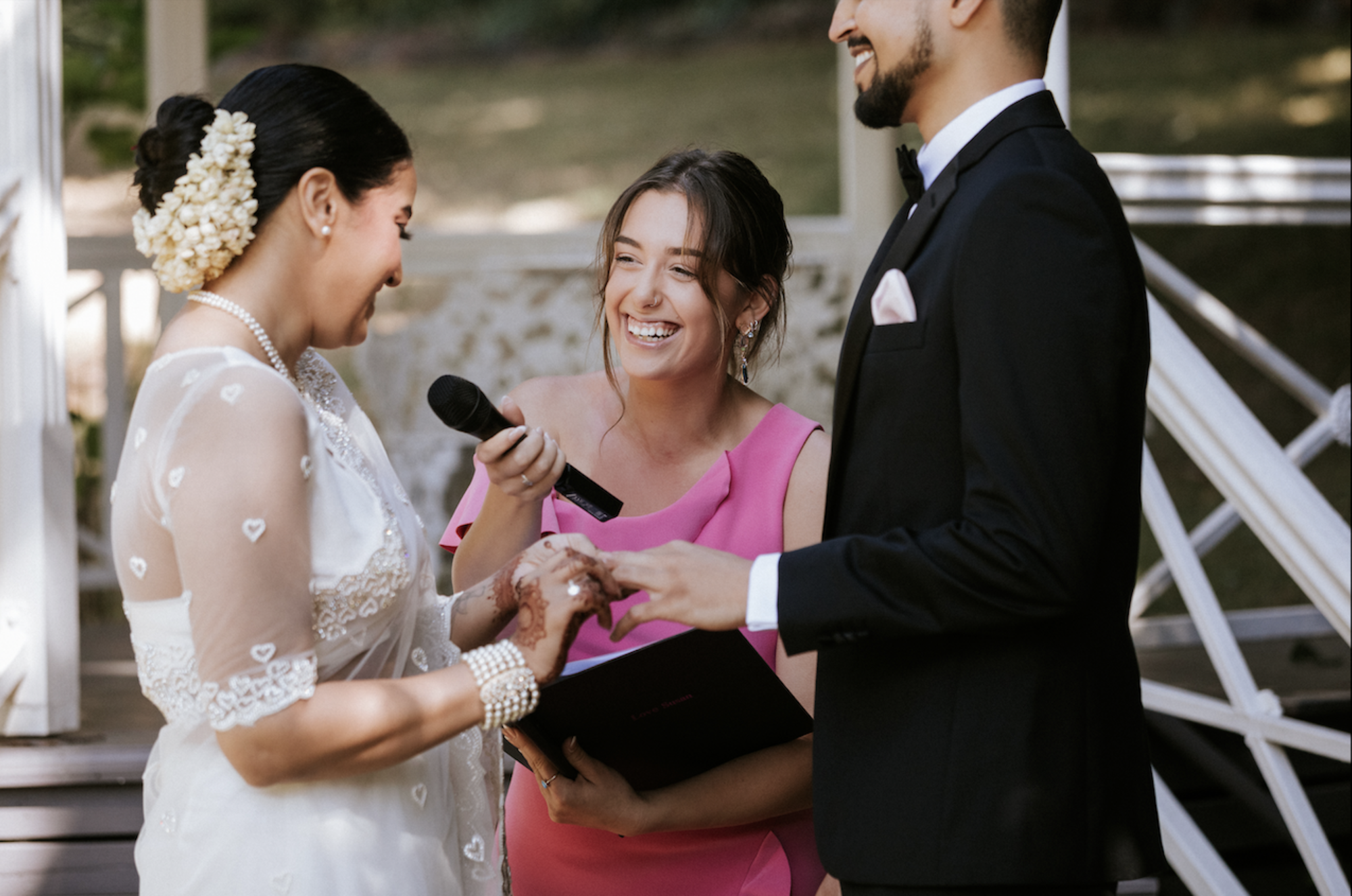 Smiling couple exchange rings during an outdoor wedding ceremony with an officiant holding a microphone.