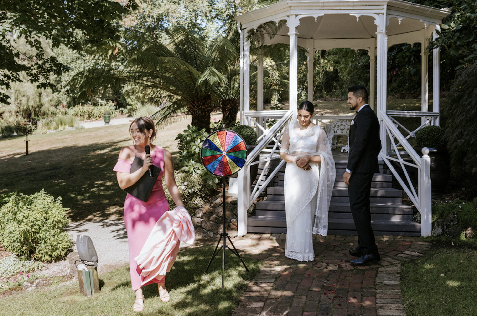 Bride and groom stand by a white garden gazebo while a celebrant leads a fun game with a colorful prize wheel.