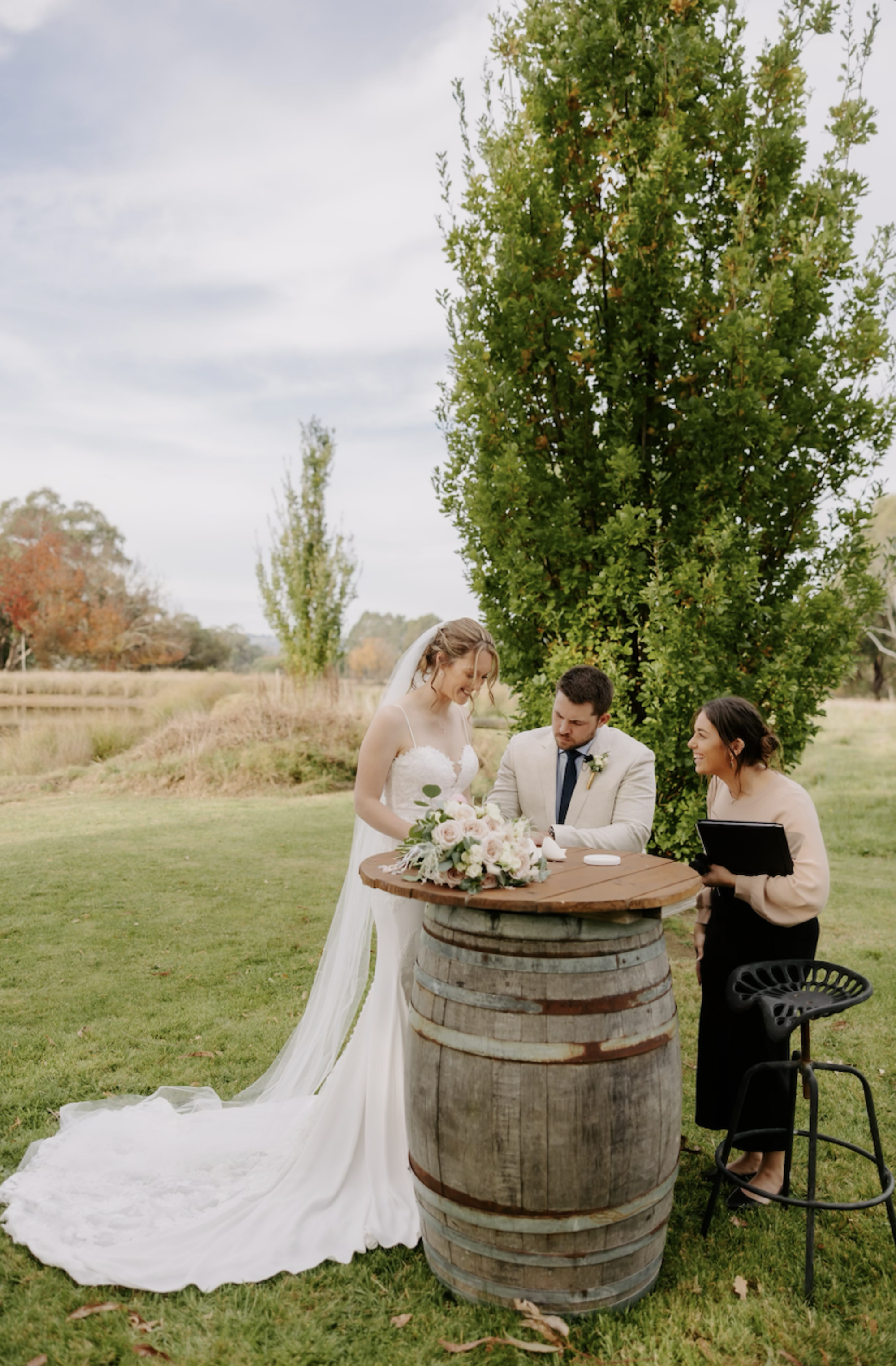 Bride and groom sign their marriage certificate at a rustic barrel table with a celebrant in a scenic outdoor garden.