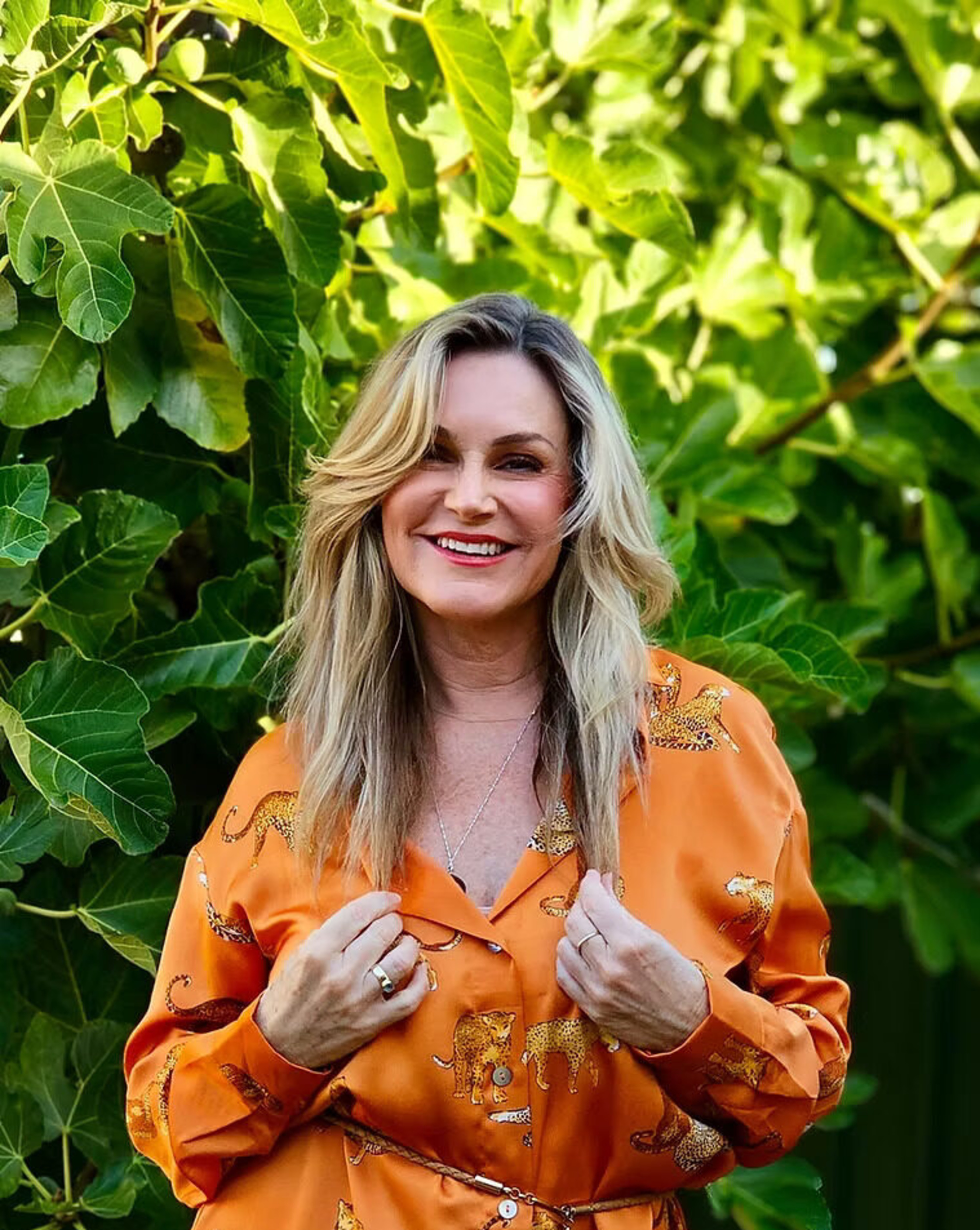 Smiling woman in an orange patterned blouse stands in front of lush green foliage.