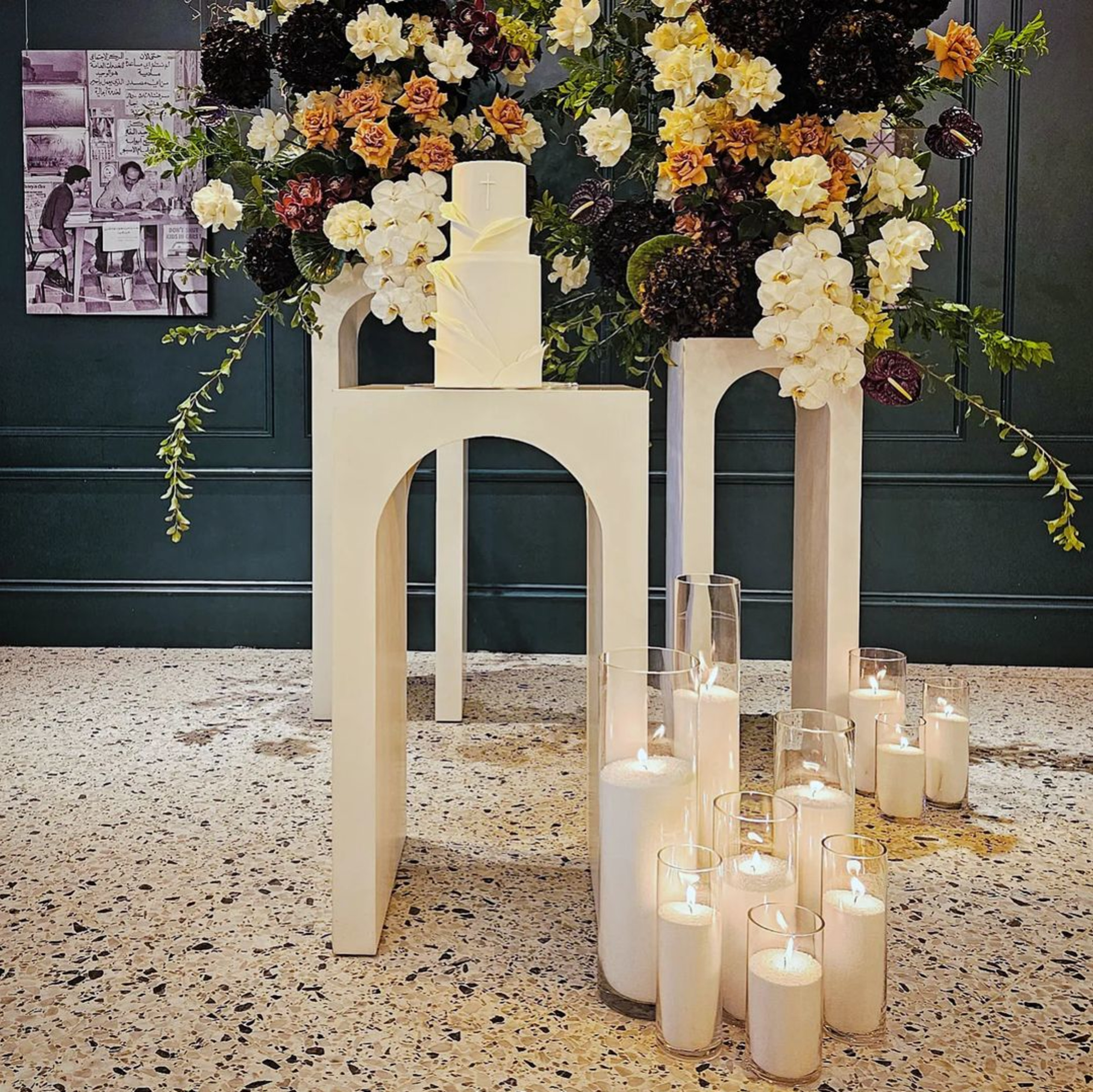 Modern wedding cake display with tall floral arrangements and glowing pillar candles on minimalist stands.