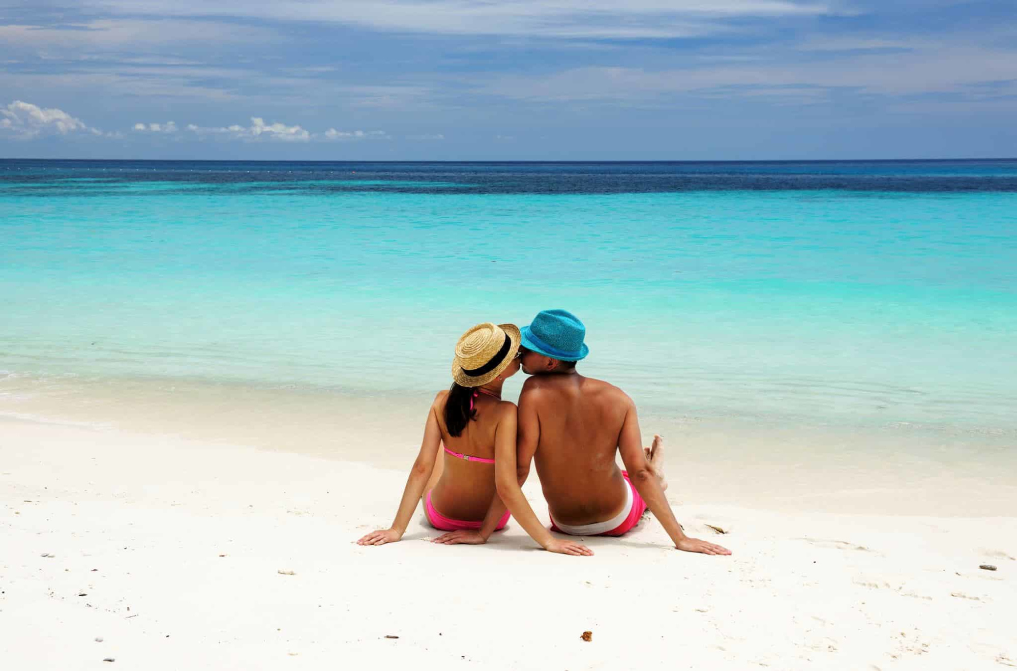 A couple in swimwear sits kissing on a white sand beach facing a turquoise ocean.