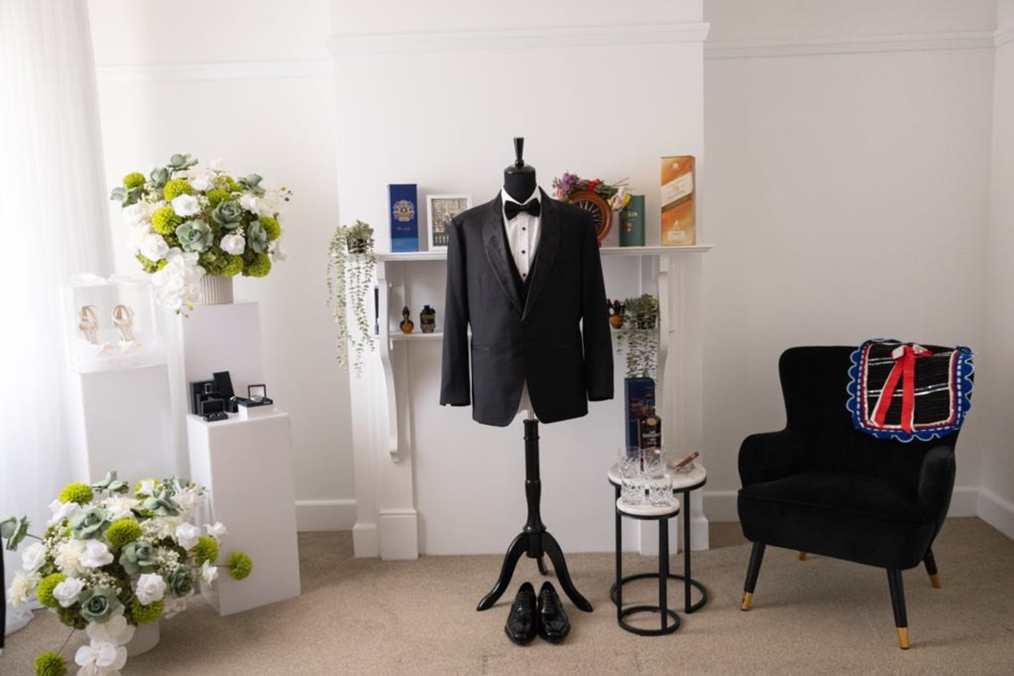 Black tuxedo on a mannequin surrounded by groom accessories and floral wedding decor in a bright room.