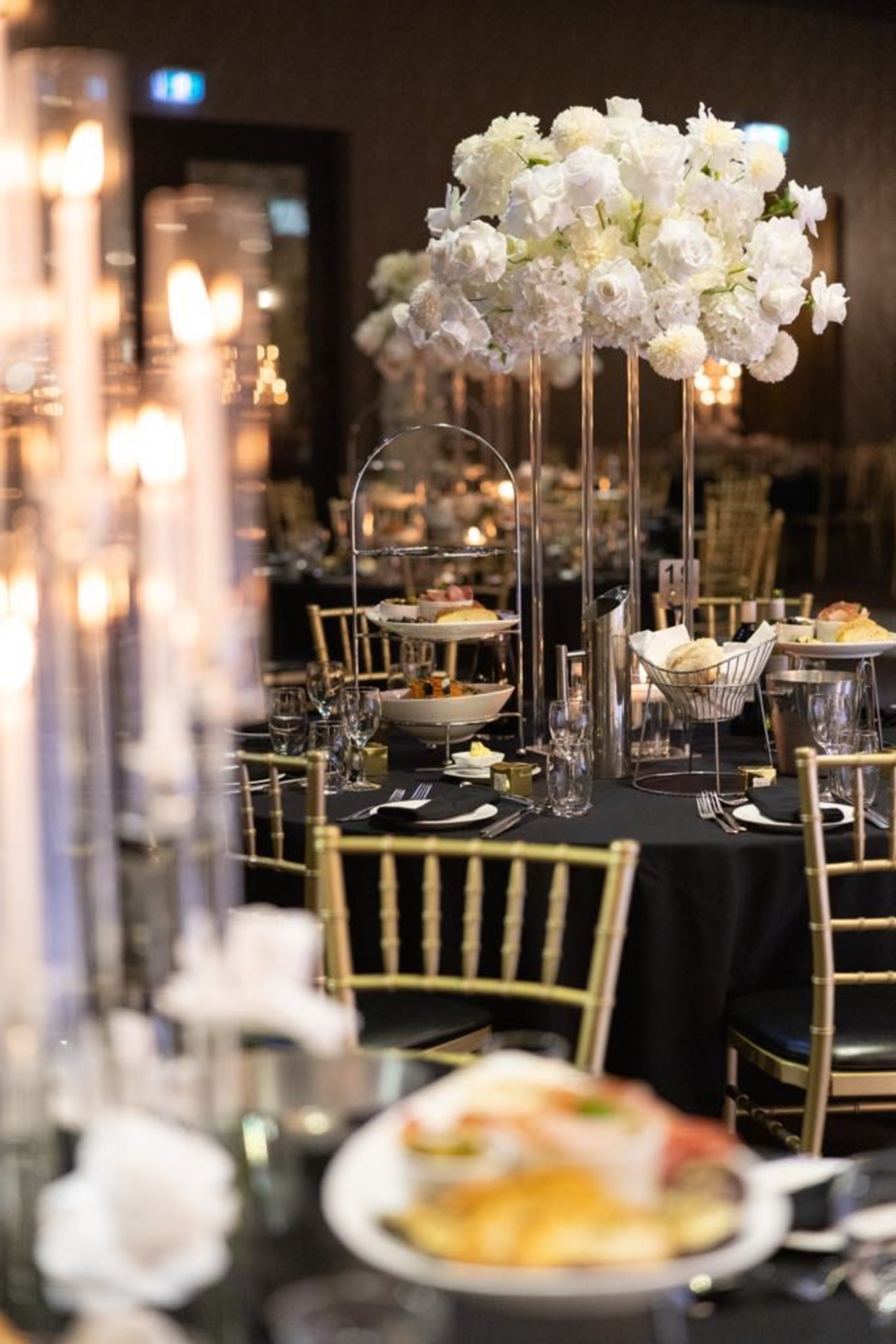 Elegant wedding reception table with tall white floral centerpiece, candlelight, and gold chairs set for a formal dinner.