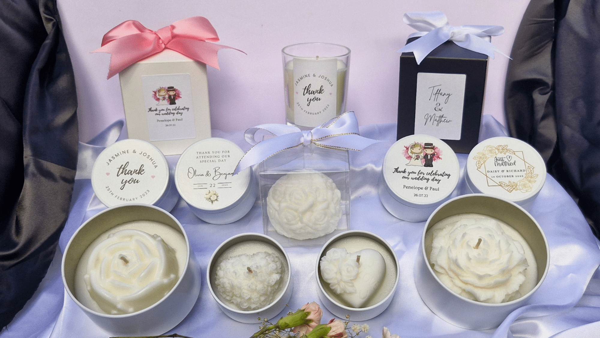 Assorted personalized wedding favor candles with floral and heart designs arranged on satin fabric.