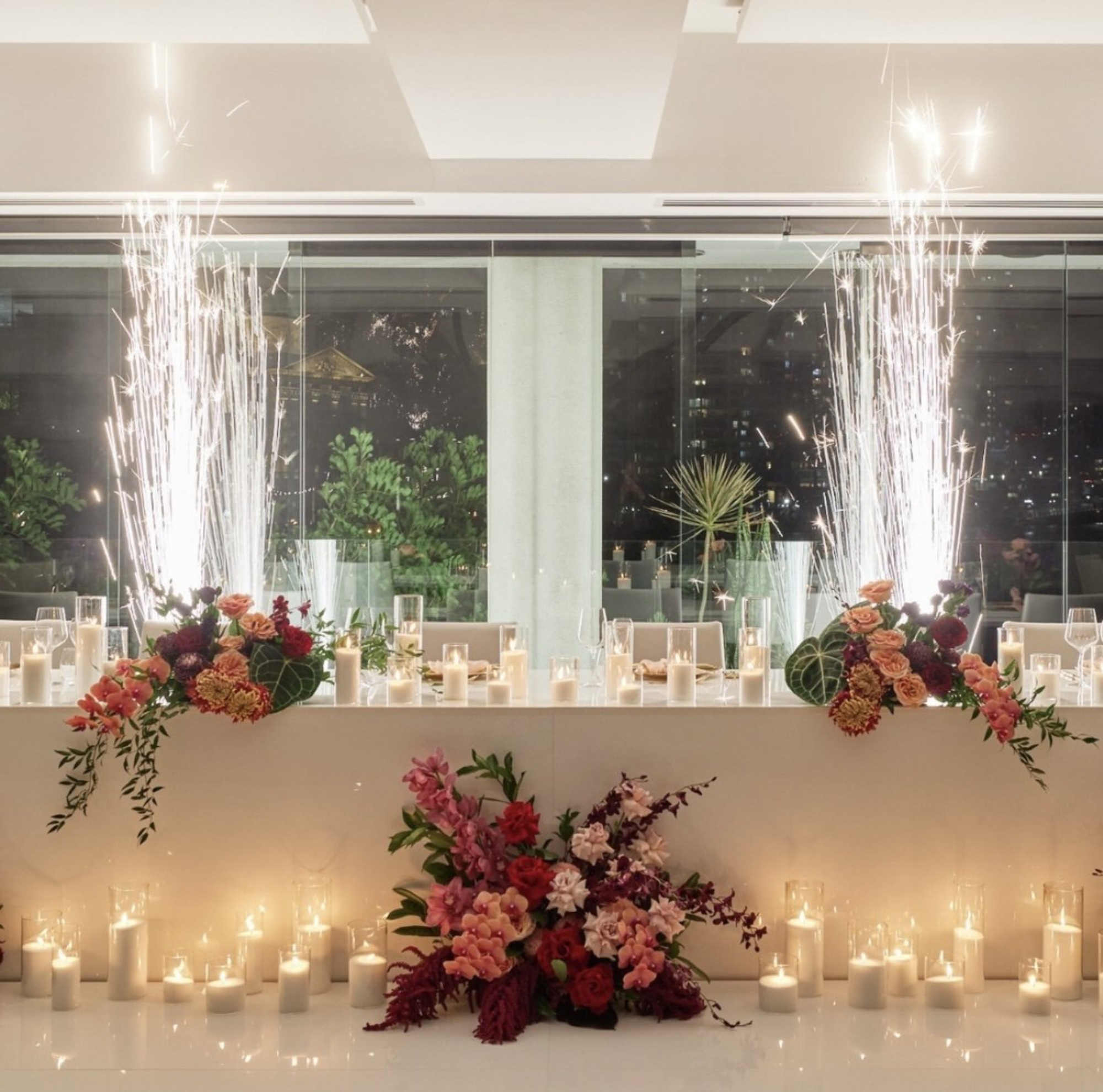 Luxurious wedding head table with candles, vibrant florals, and indoor sparklers against a nighttime window backdrop.