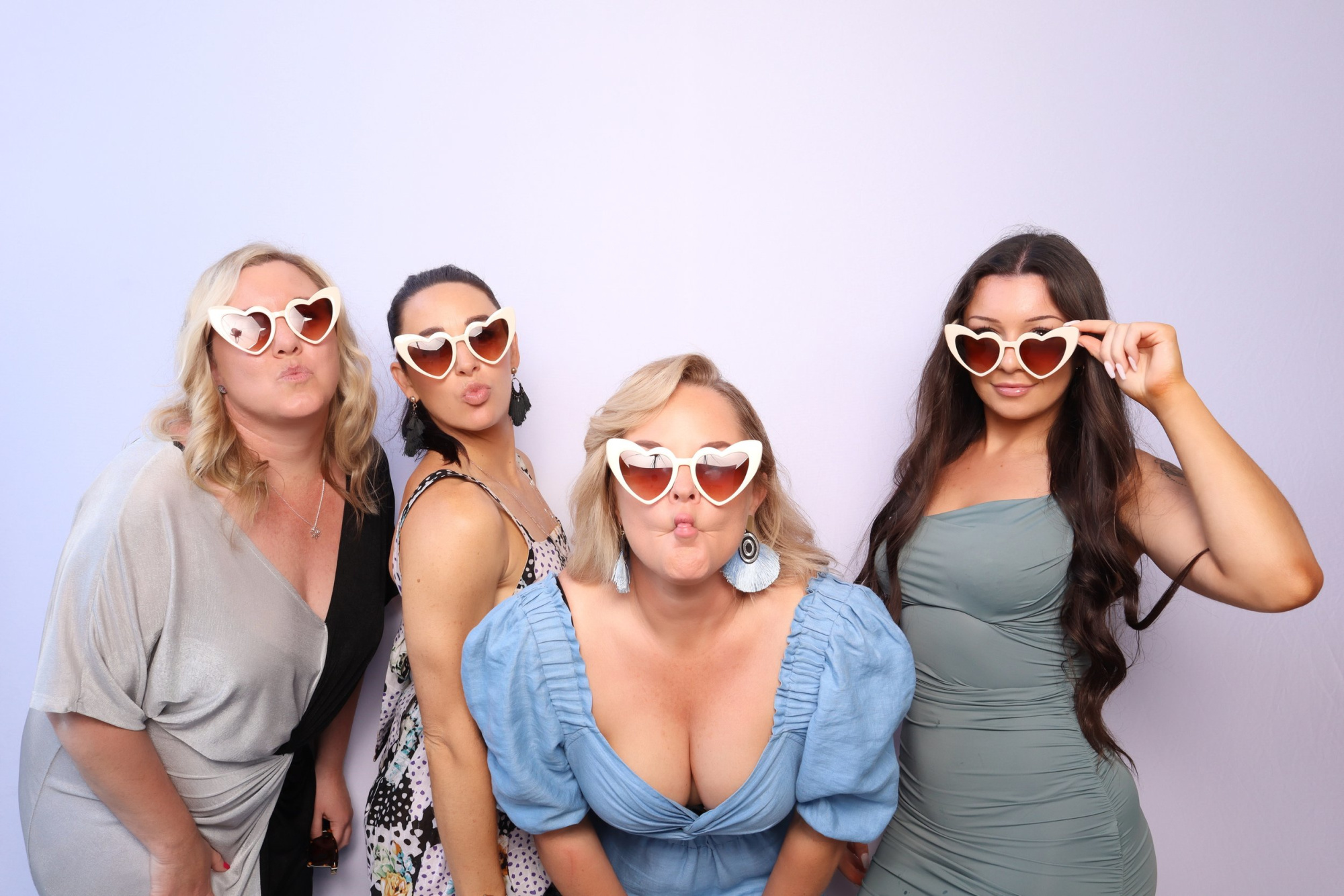 Four women in heart-shaped sunglasses pose playfully in a wedding photo booth against a light backdrop.