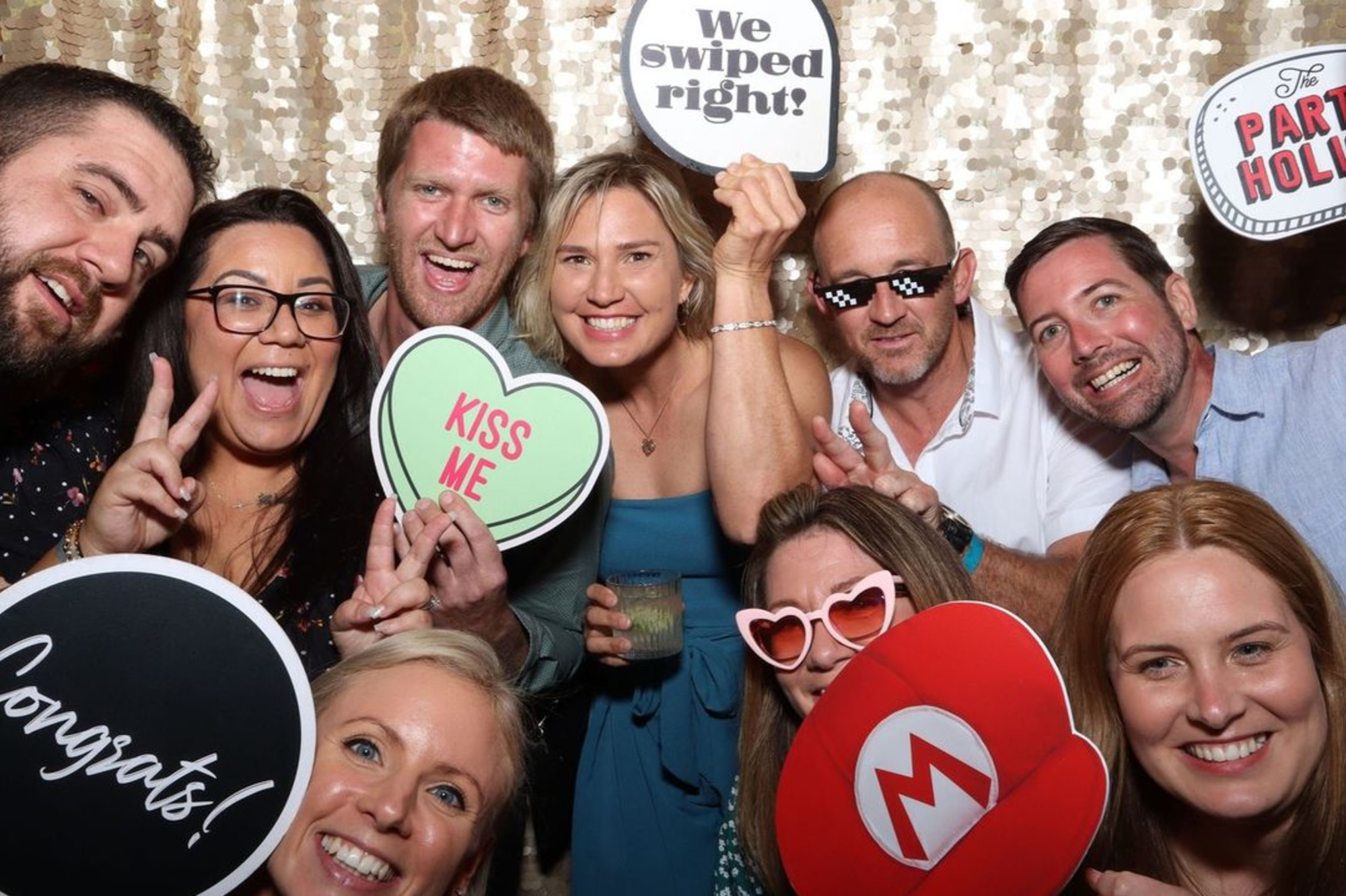 Group of smiling wedding guests posing with playful photo booth props in front of a sequin backdrop.
