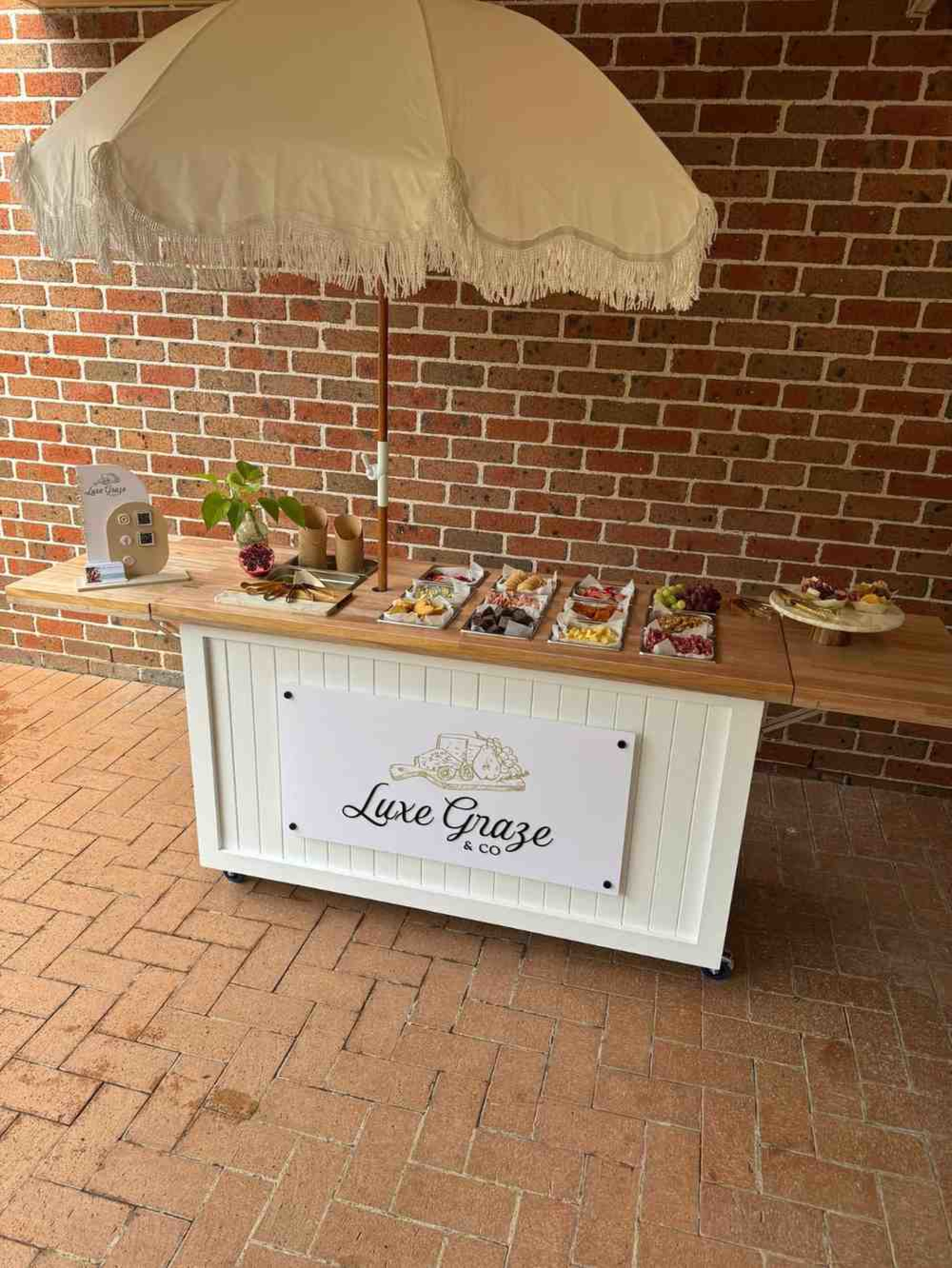 Outdoor grazing station with Luxe Graze & Co sign, umbrella, and assorted wedding canapés on display.