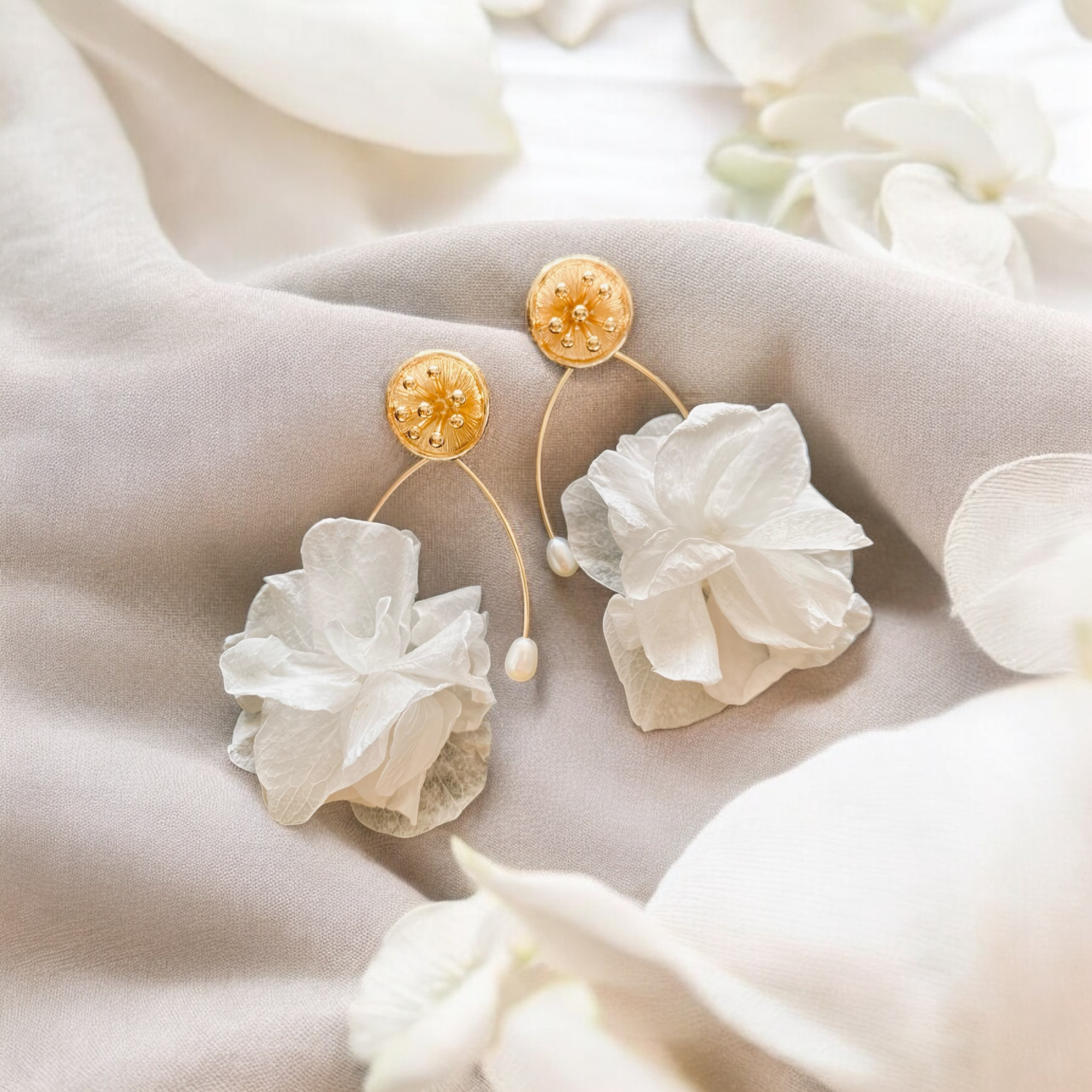 Gold floral bridal earrings with white petals and pearls styled on soft neutral fabric.