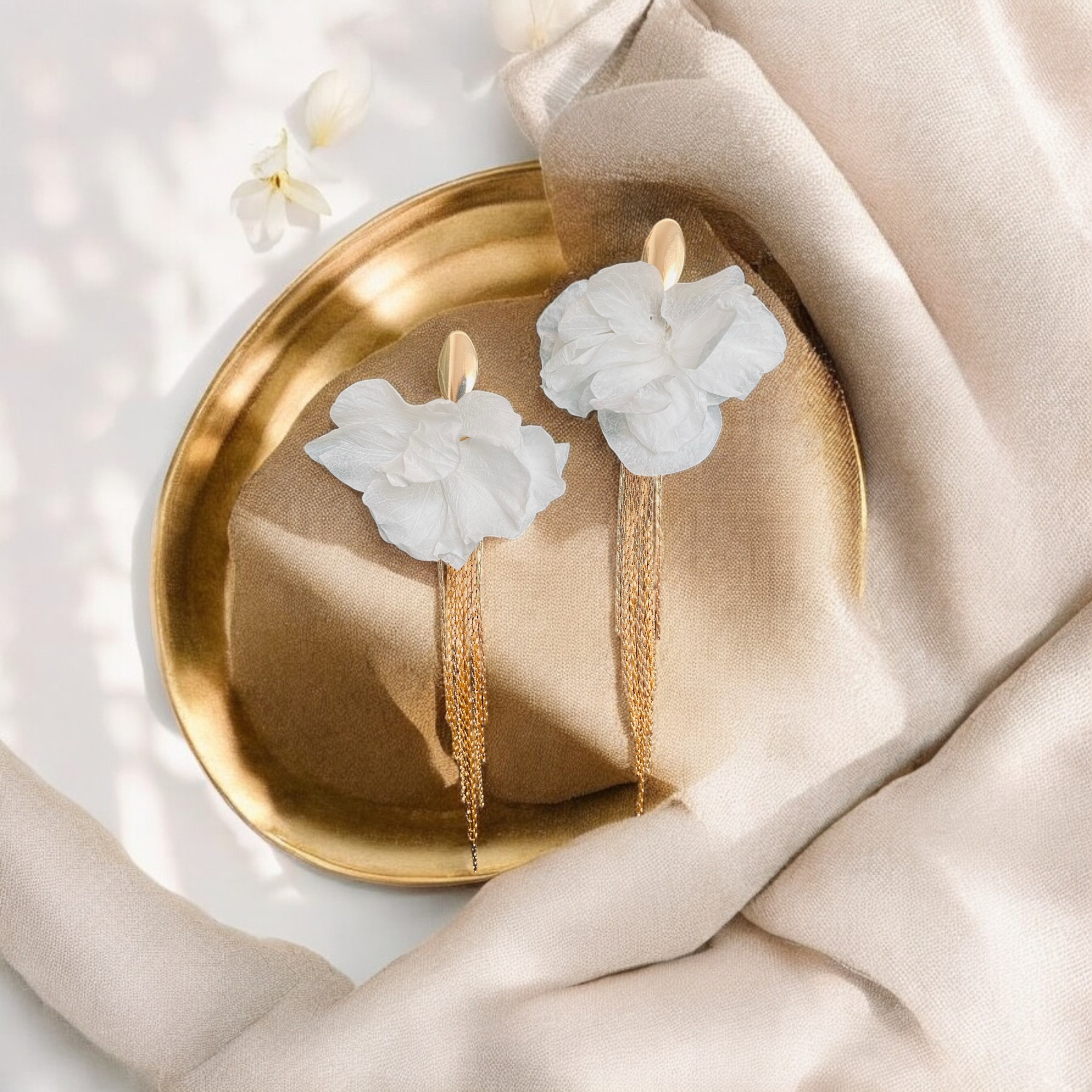 Gold chain earrings with white floral details styled on a gold plate and soft beige fabric.