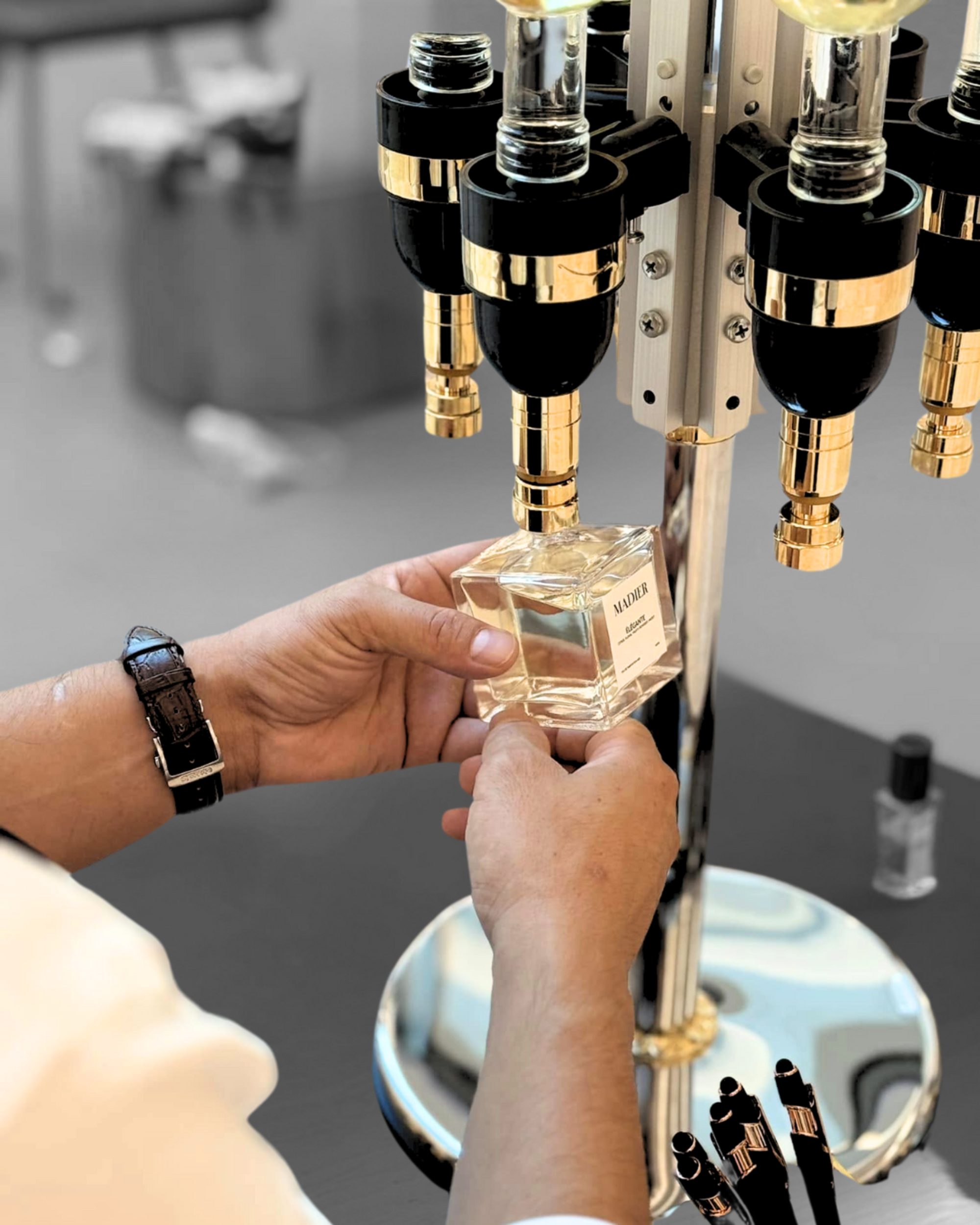 Hands holding a perfume bottle beneath a gold and black fragrance dispensing machine.