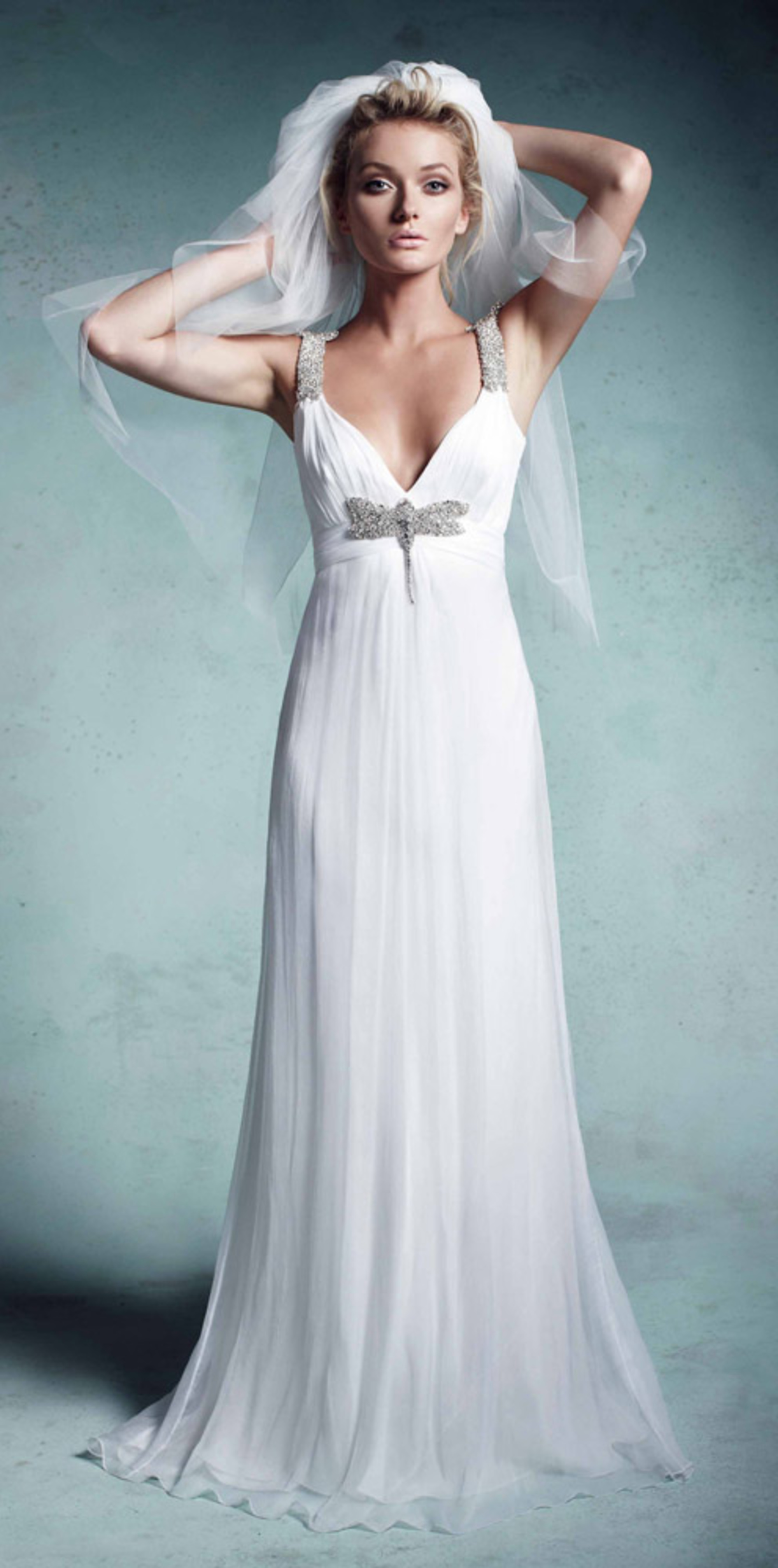 Bride wearing a simple white wedding gown with jeweled straps and veil against a soft blue backdrop.