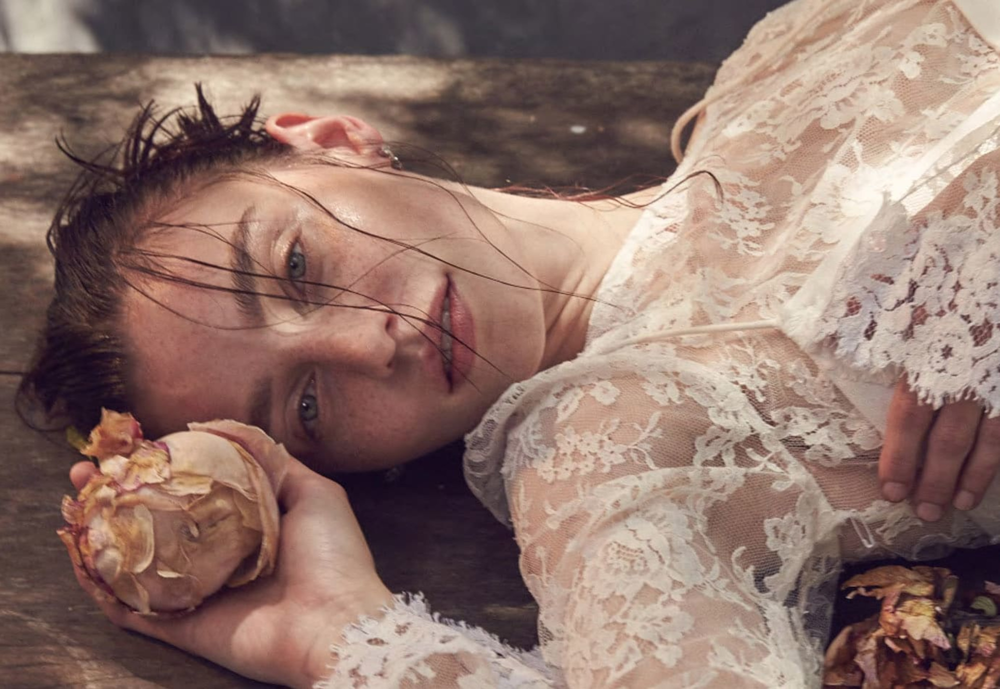 Dreamy bridal portrait of a woman in a lace gown lying on a wooden surface holding a faded flower.