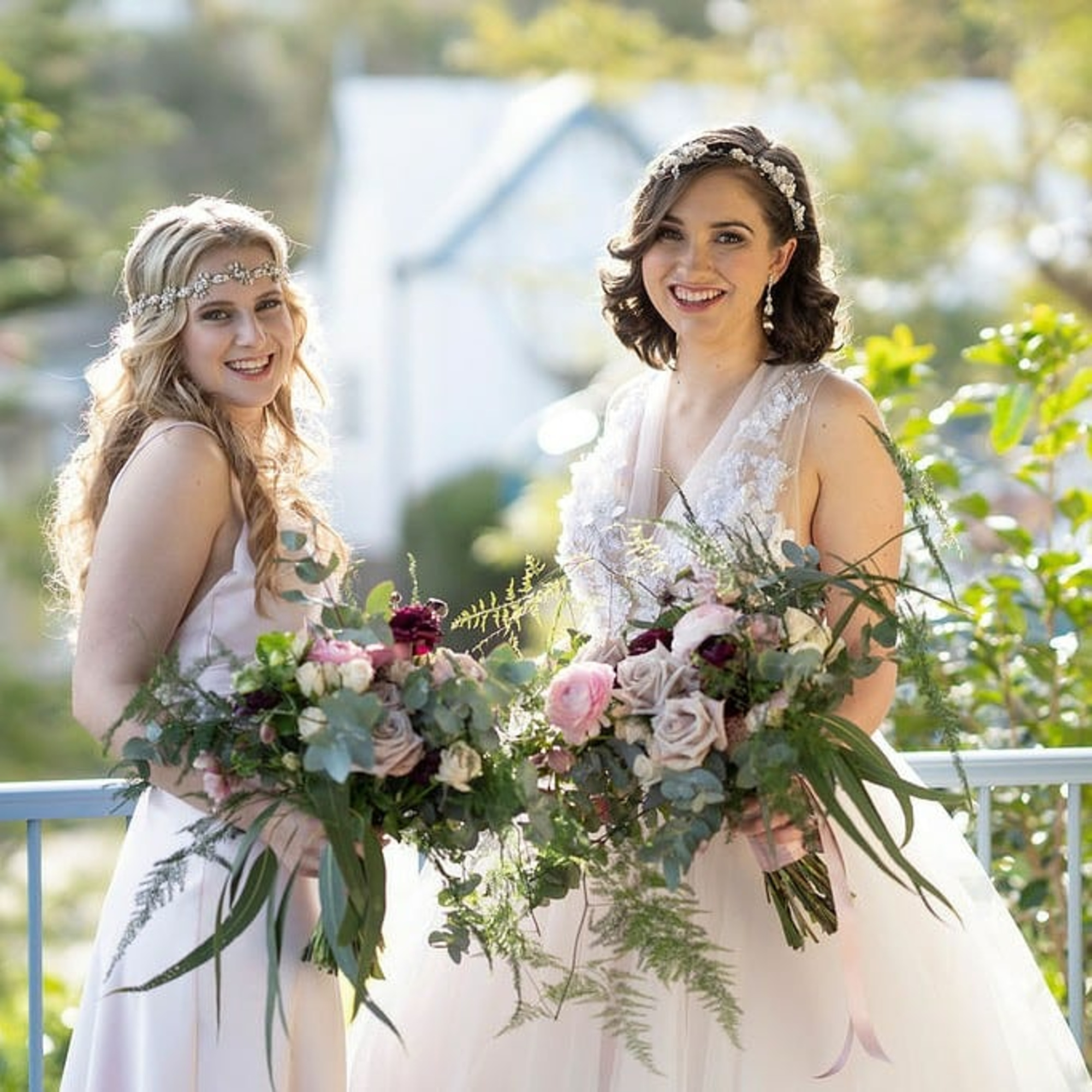 Two brides in light gowns hold lush floral bouquets while posing outdoors in soft natural light.