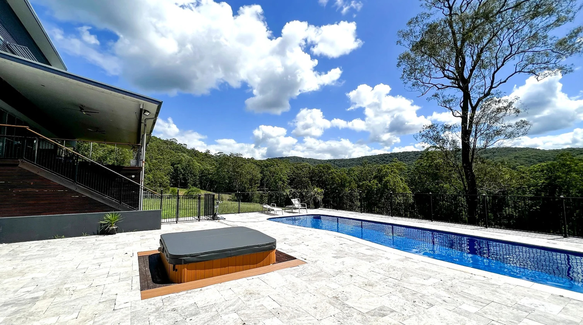 Modern estate poolside terrace with hot tub overlooking forested hills under a bright blue sky.