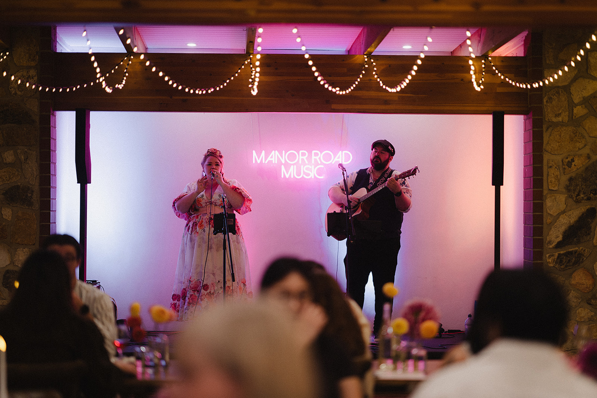 Acoustic duo performs on stage under a neon Manor Road Music sign at a wedding reception.