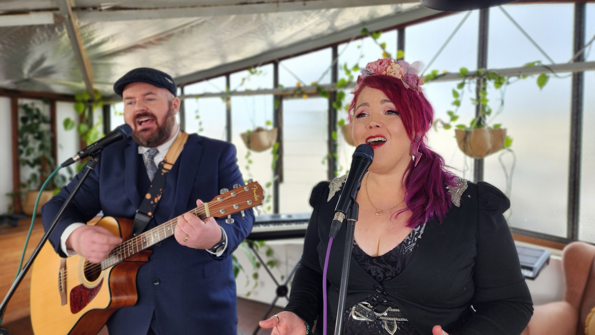Male guitarist and female vocalist performing live music together in a bright, plant-filled indoor venue.