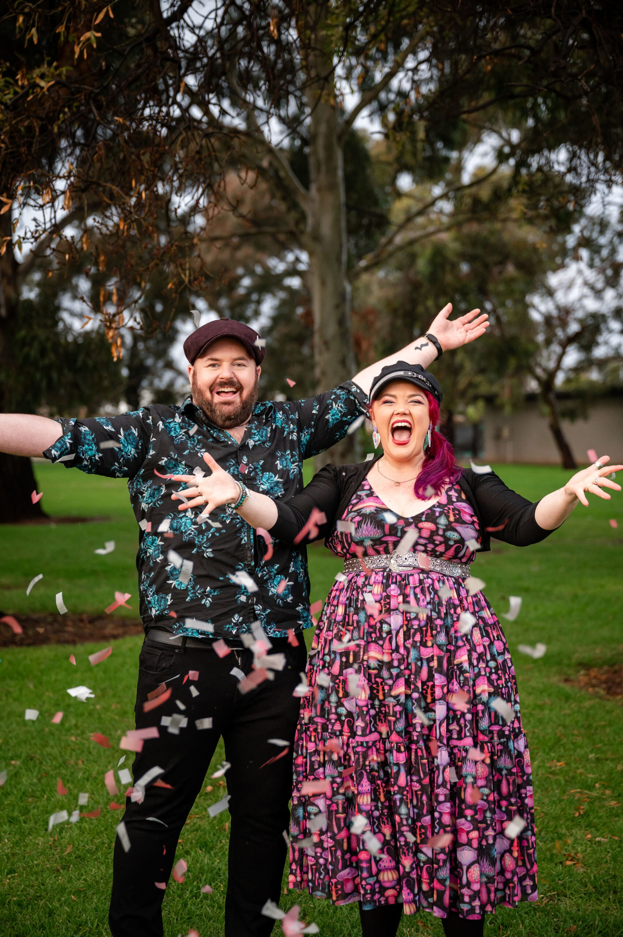 Joyful couple in colorful outfits throwing confetti in a green park setting.