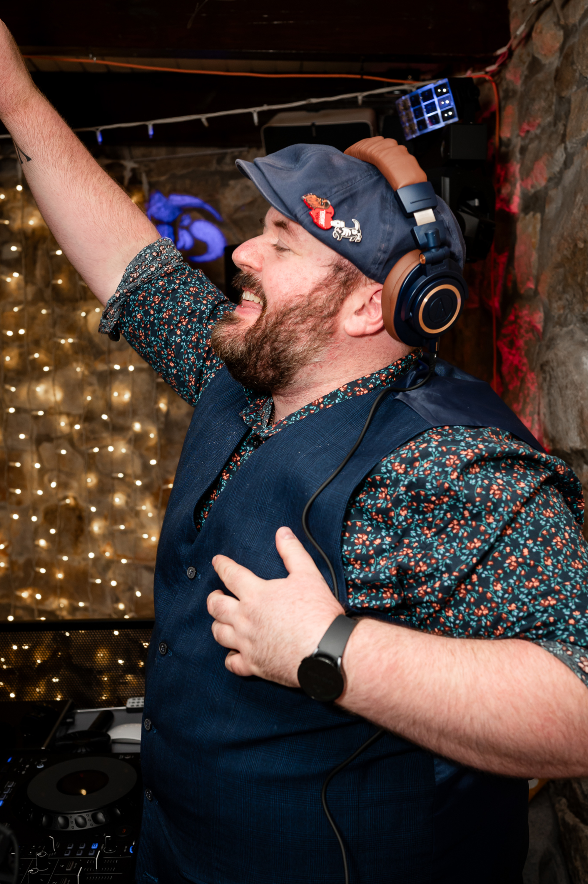 Wedding DJ in headphones energetically mixing music at a lively reception with twinkling lights behind.