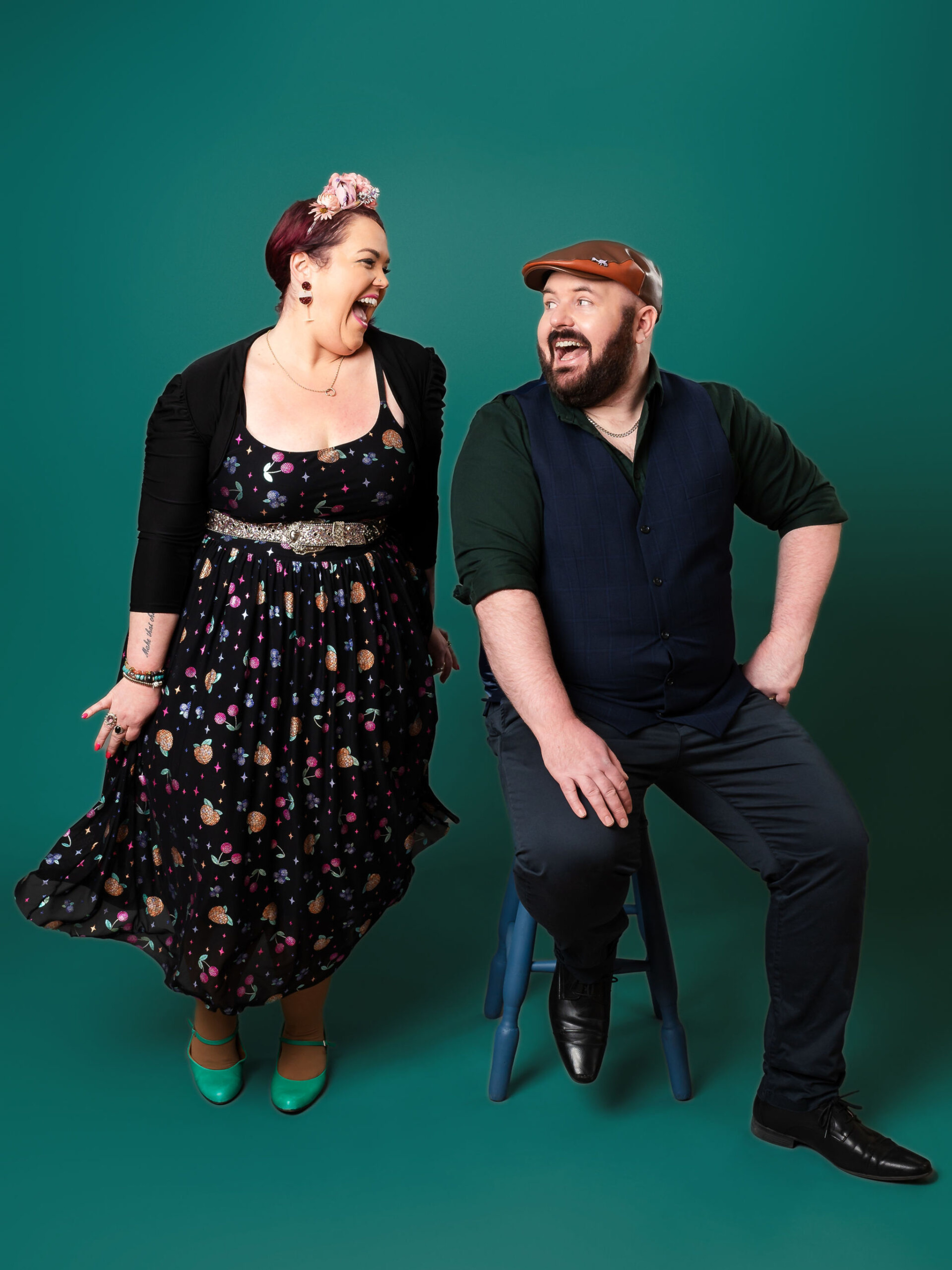 Smiling wedding vendor duo posing playfully together against a solid teal backdrop.