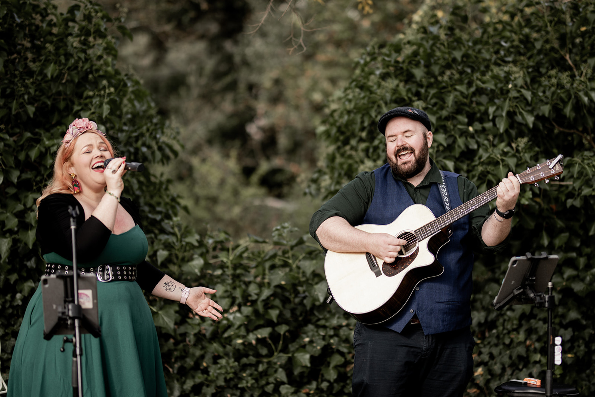 Acoustic duo singing and playing guitar at an outdoor wedding surrounded by greenery.