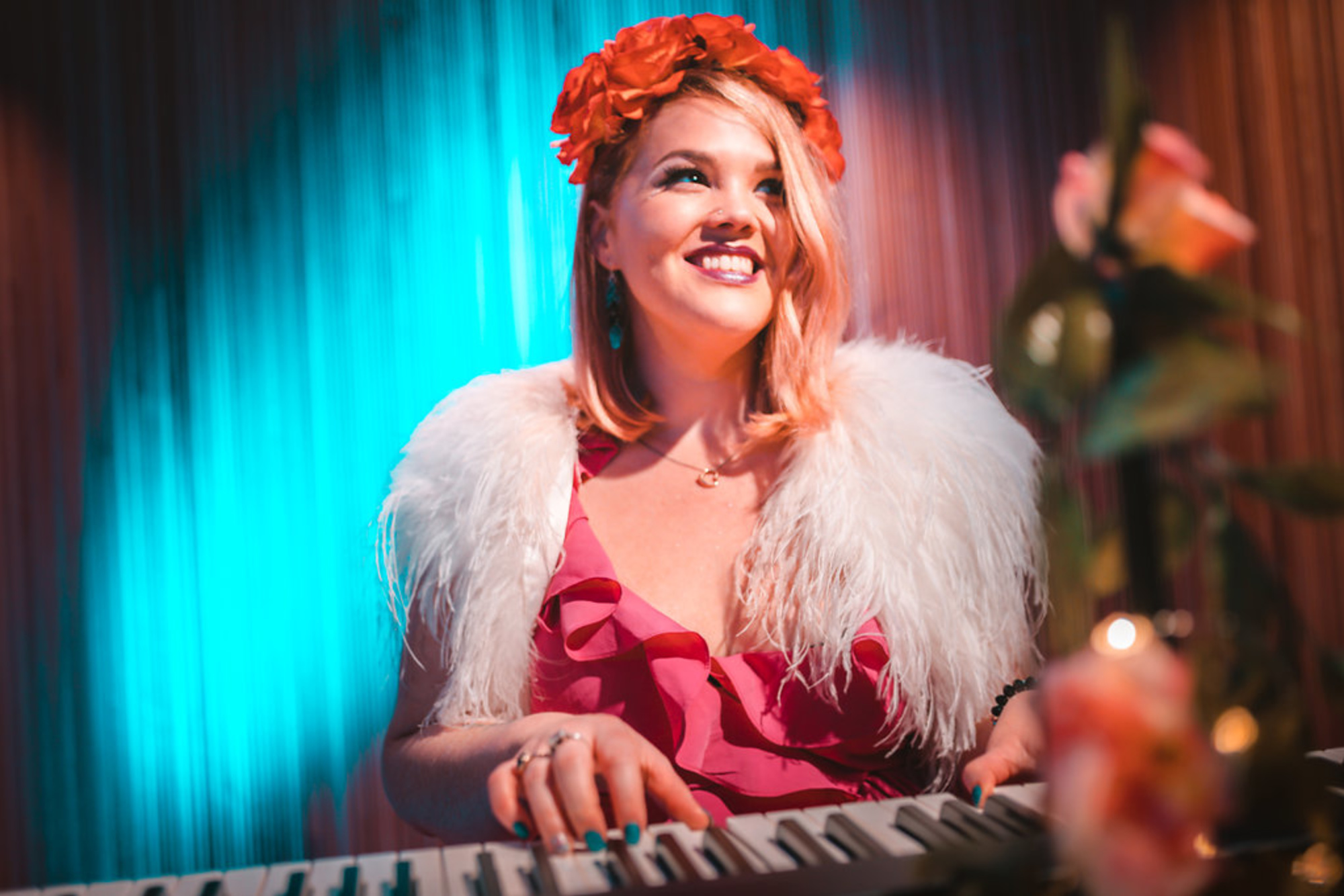Smiling wedding pianist in a floral headpiece and feathered shawl playing keyboard under colorful lighting.