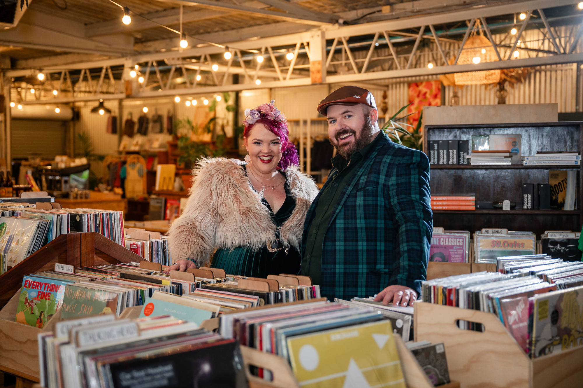 Smiling couple pose among vinyl record bins in a cozy, eclectic indoor shop with warm string lights.