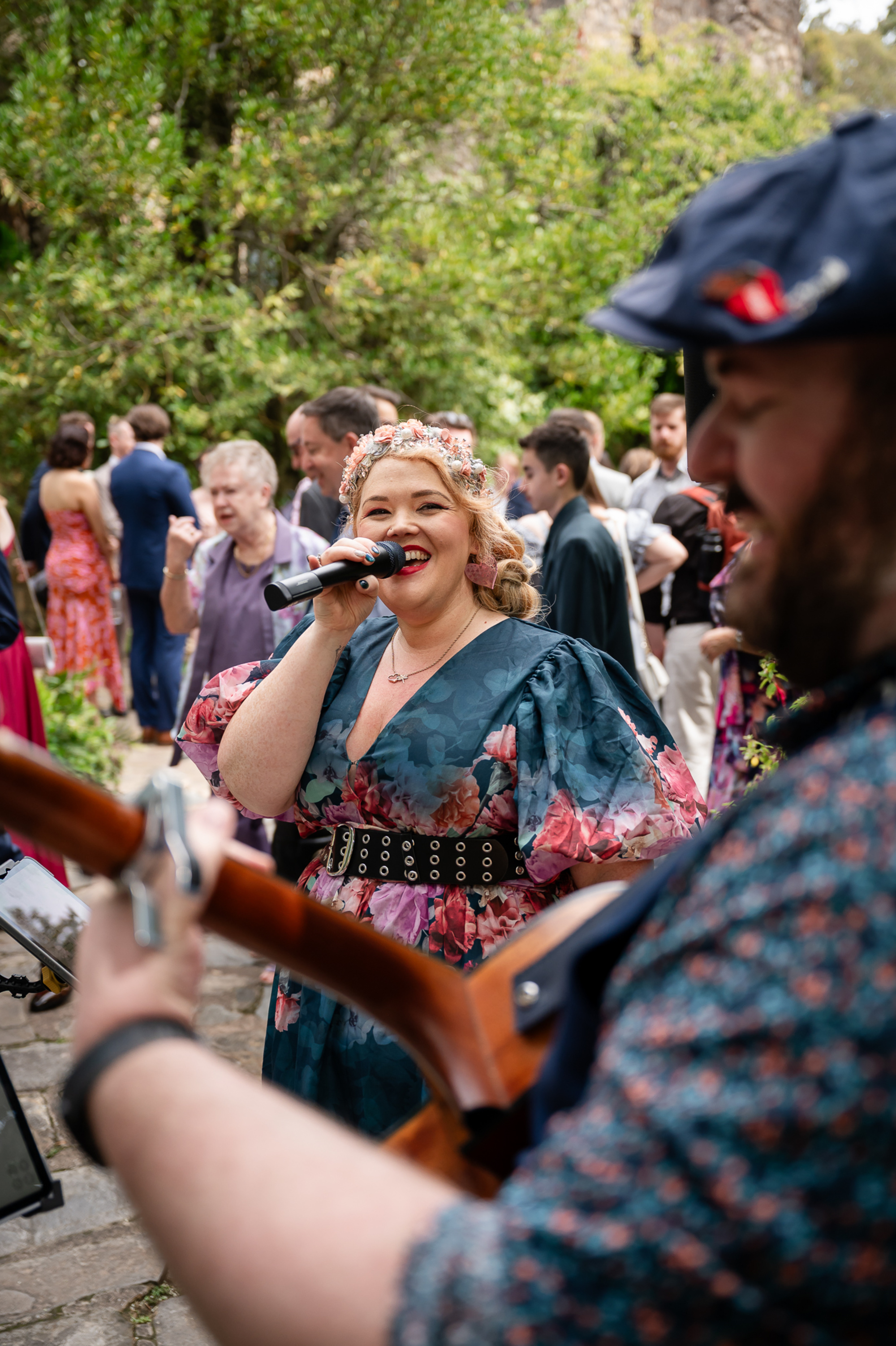 Smiling wedding singer performs with guitarist at an outdoor reception surrounded by guests.