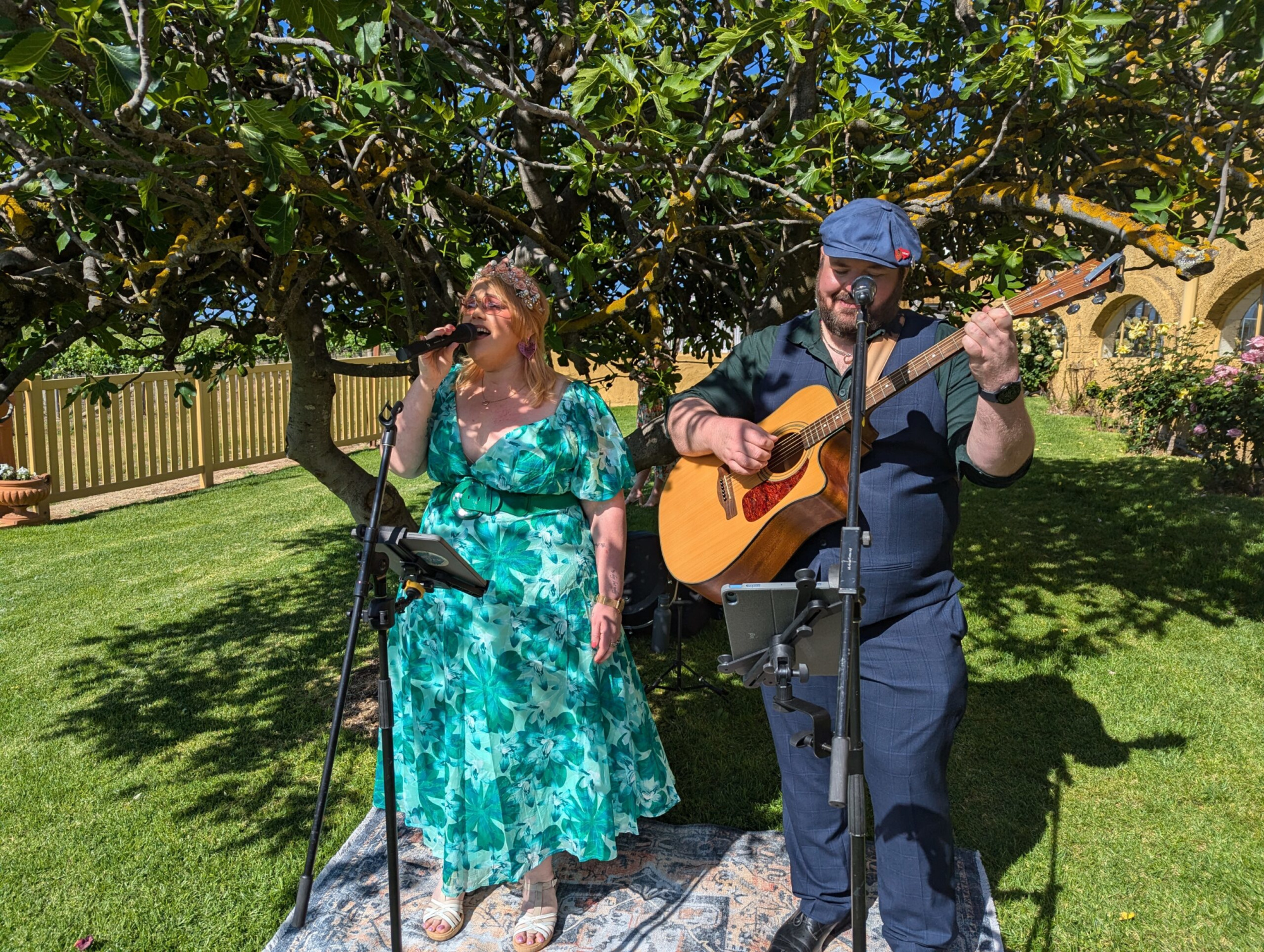 Acoustic singer and guitarist perform together under a tree at an outdoor garden wedding.