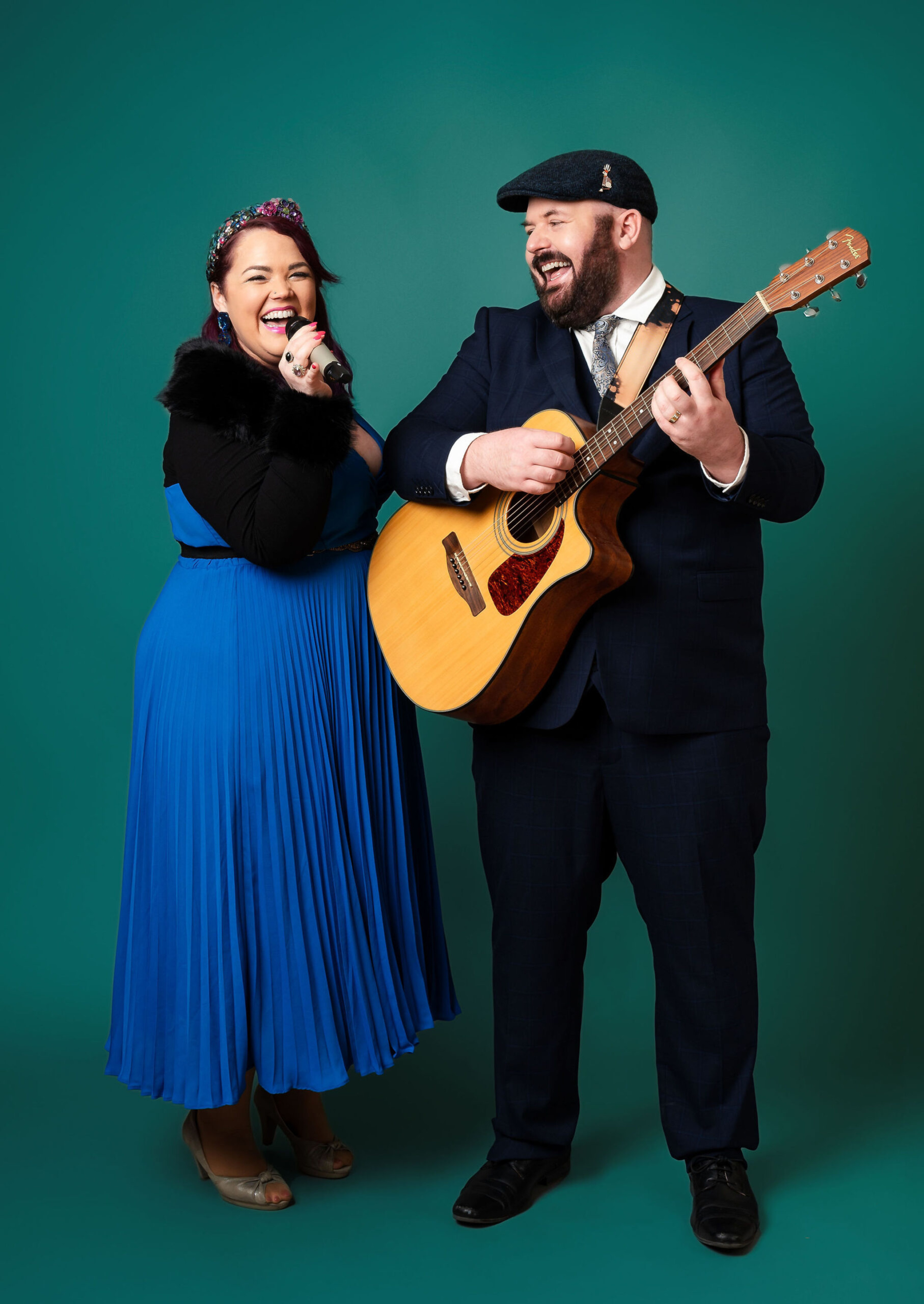 Smiling musical duo with a singer and guitarist posing against a teal backdrop for wedding entertainment.