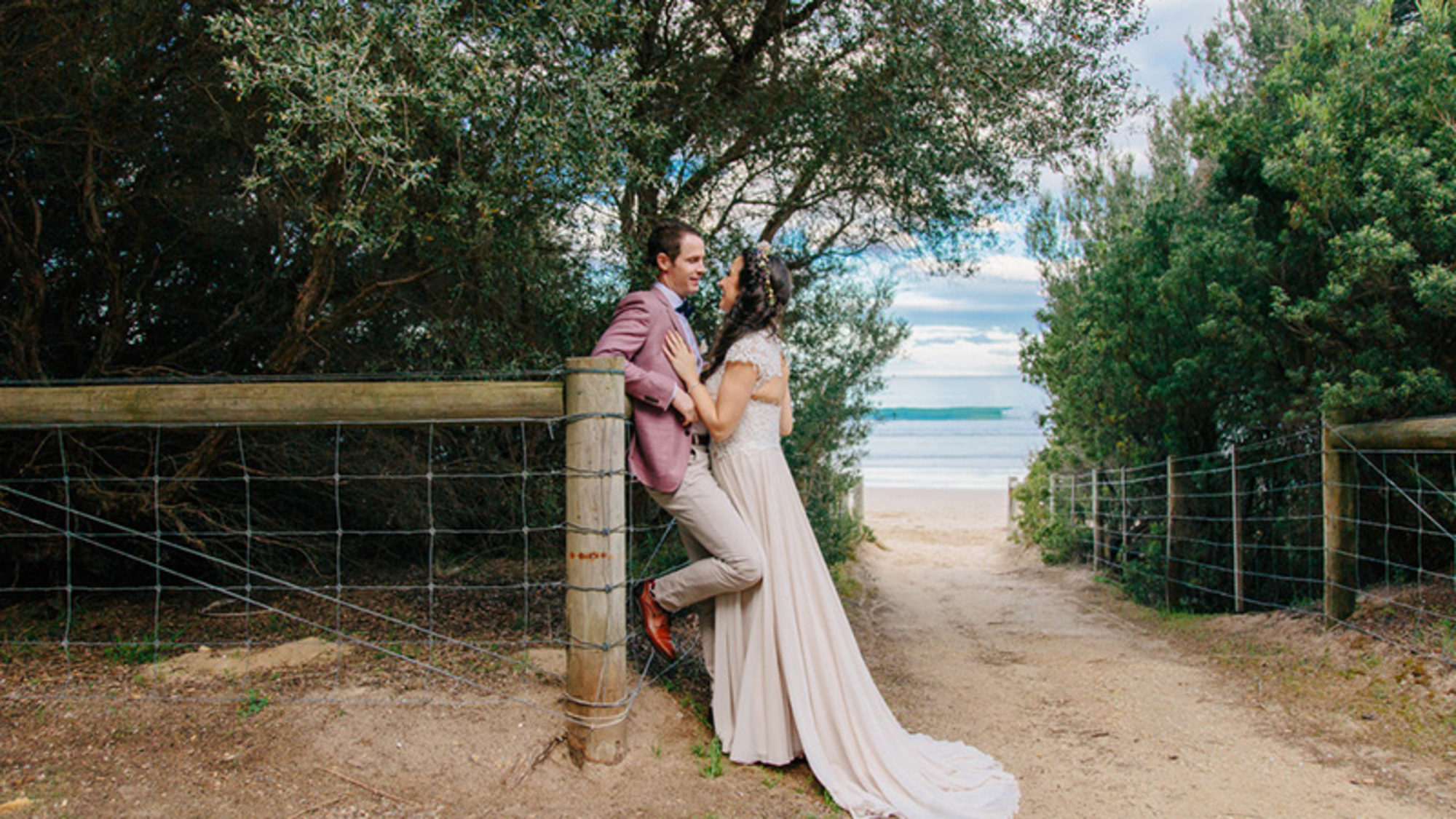 Bride and groom share a quiet moment on a sandy path between trees leading to the beach.