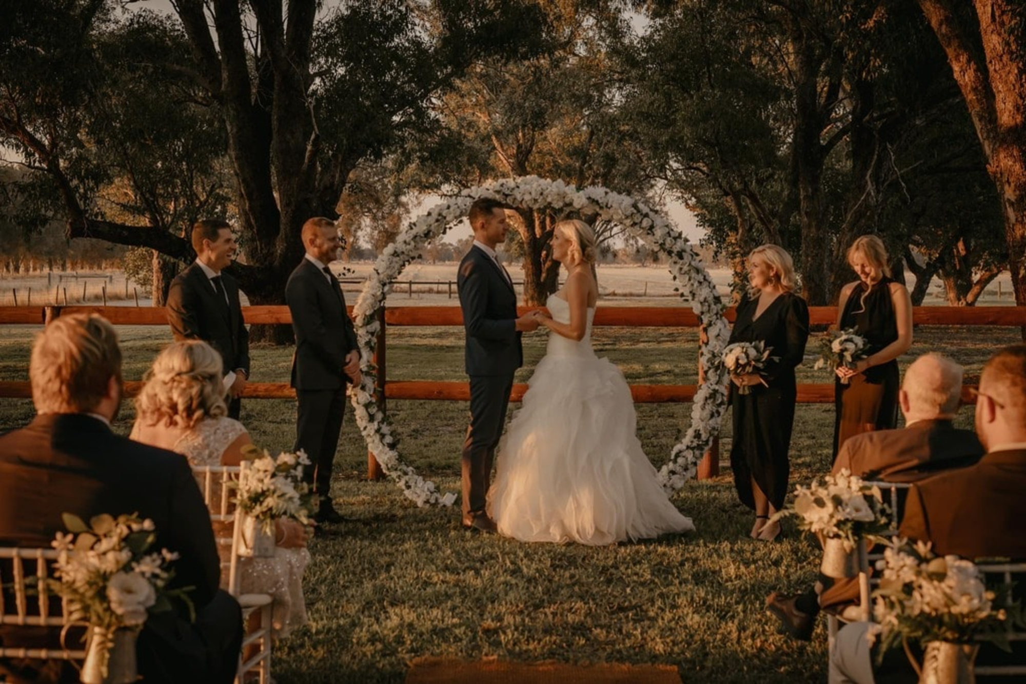 Outdoor sunset wedding ceremony with a couple exchanging vows under a circular flower arch before seated guests.