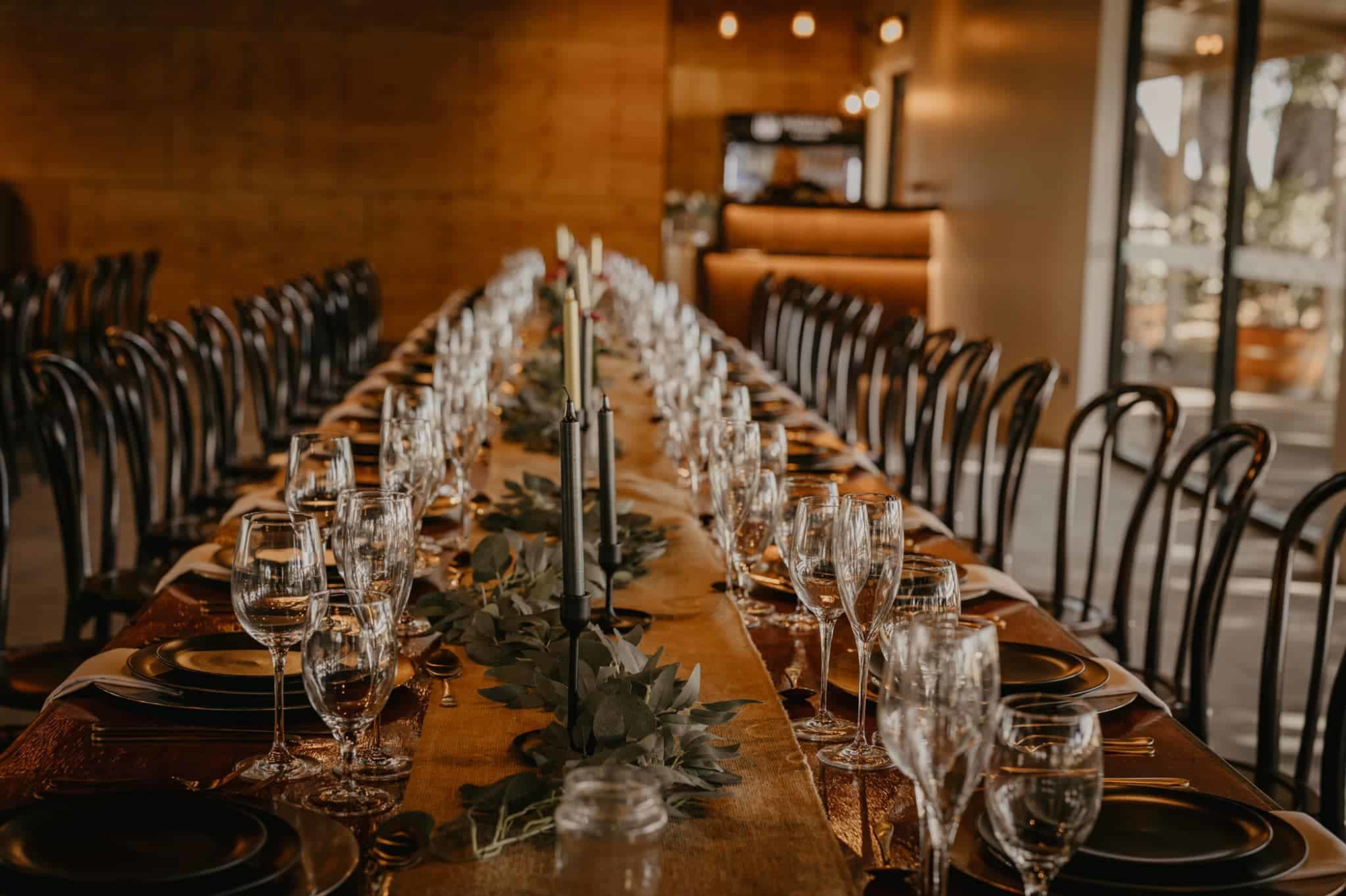 Elegant long wedding reception table set with glassware, candles, and greenery in a warm indoor venue.