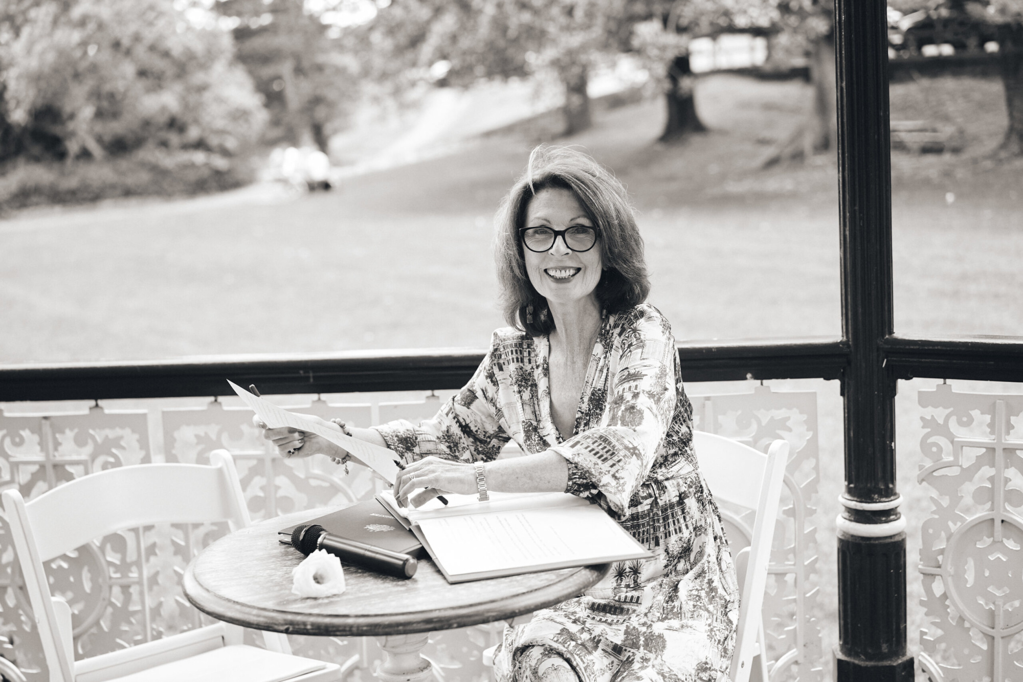 A smiling wedding celebrant sits at a small table outdoors reviewing ceremony papers with a microphone nearby.