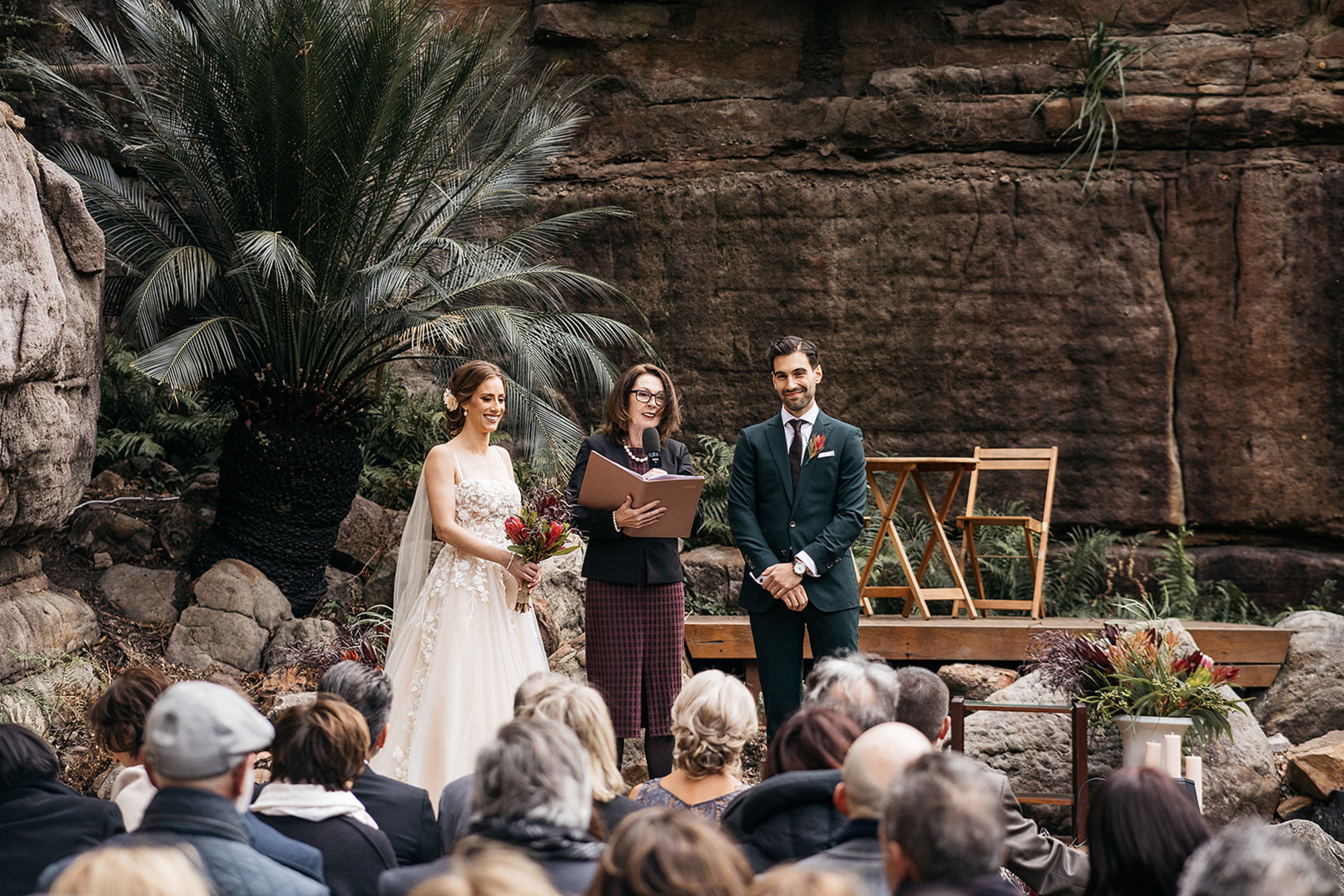Outdoor wedding ceremony with a couple and celebrant standing before guests against a rocky, lush garden backdrop.