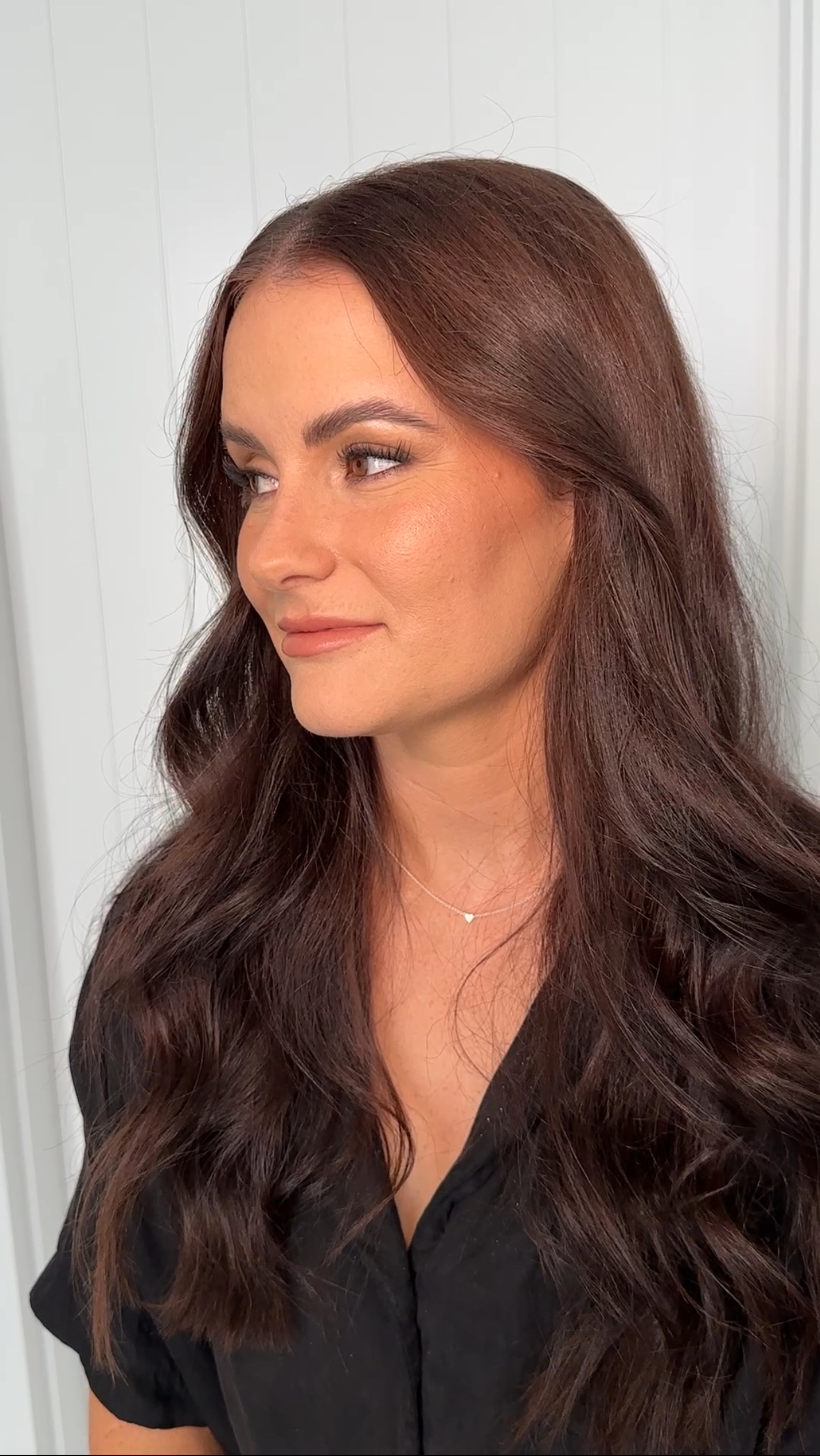 Woman with long wavy brunette hair and soft glam makeup looking to the side against a light background.