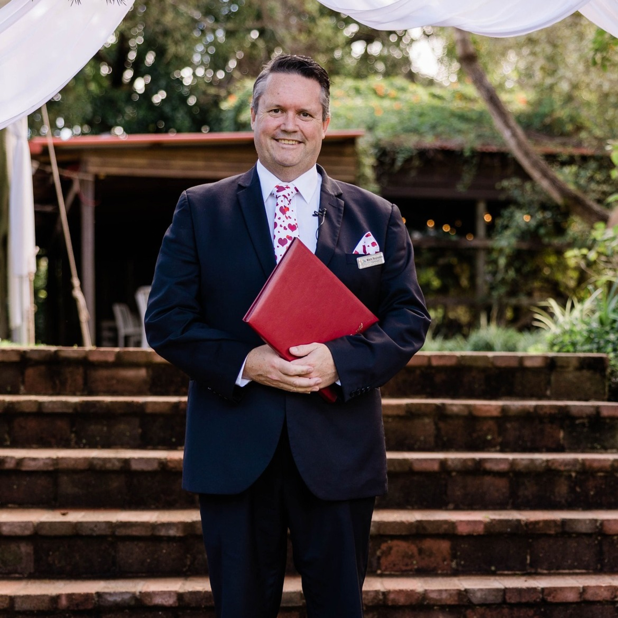Wedding officiant in a suit holding a red folder, standing on brick steps in an outdoor garden ceremony space.