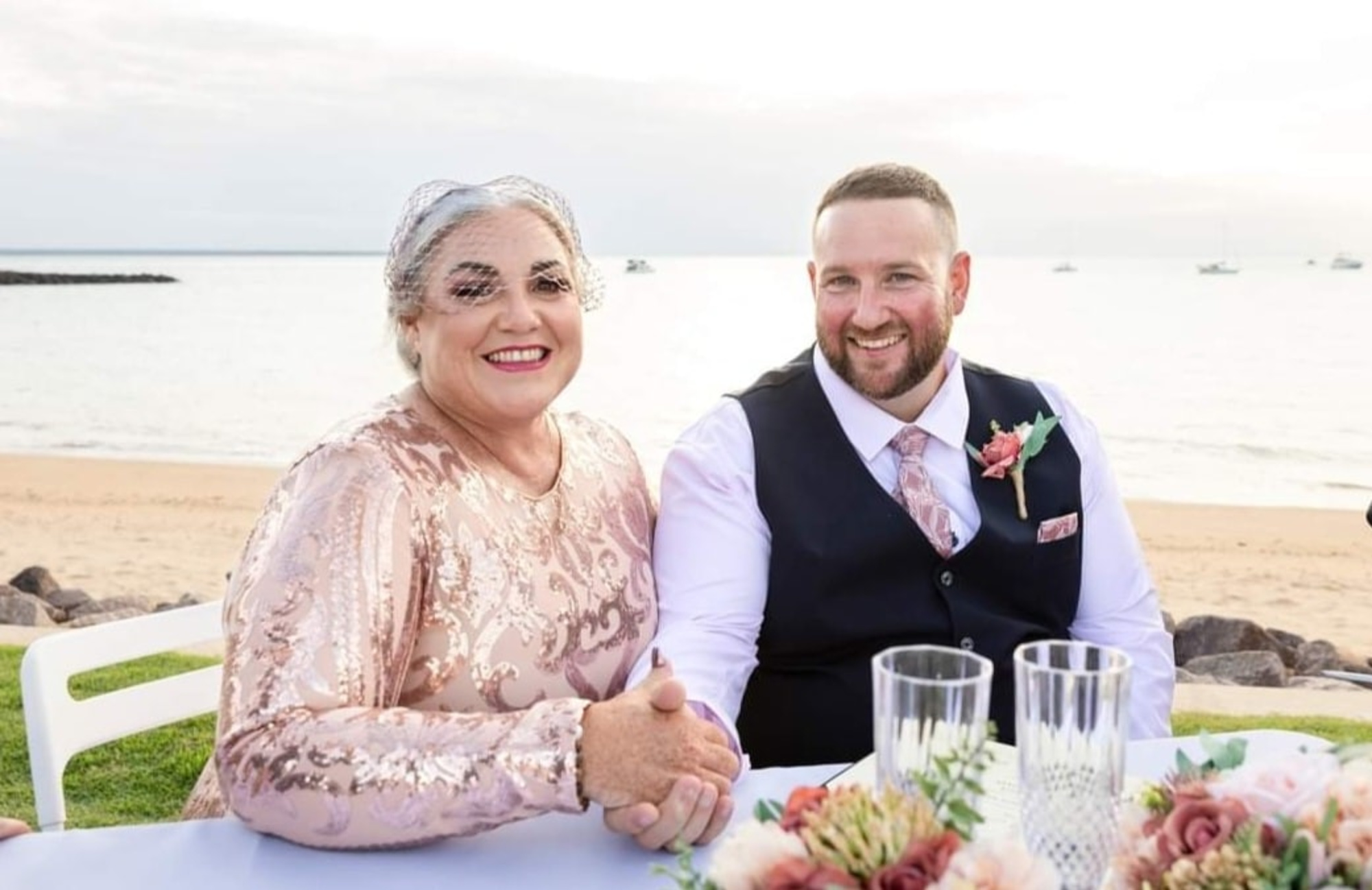 Smiling wedding couple seated at a decorated table by the beach during their seaside reception.