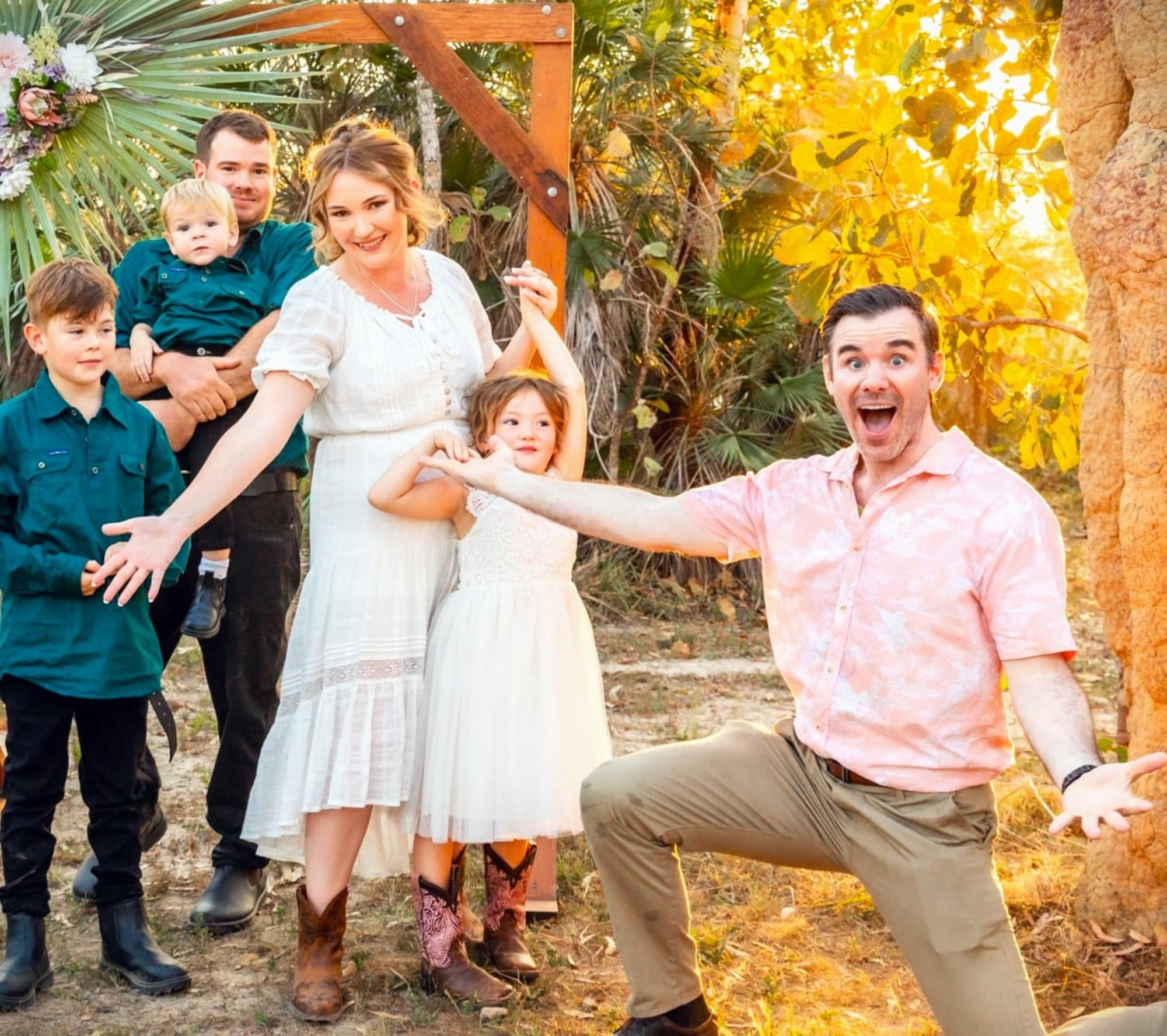 Cheerful family poses playfully under a rustic wooden arch at an outdoor wedding.