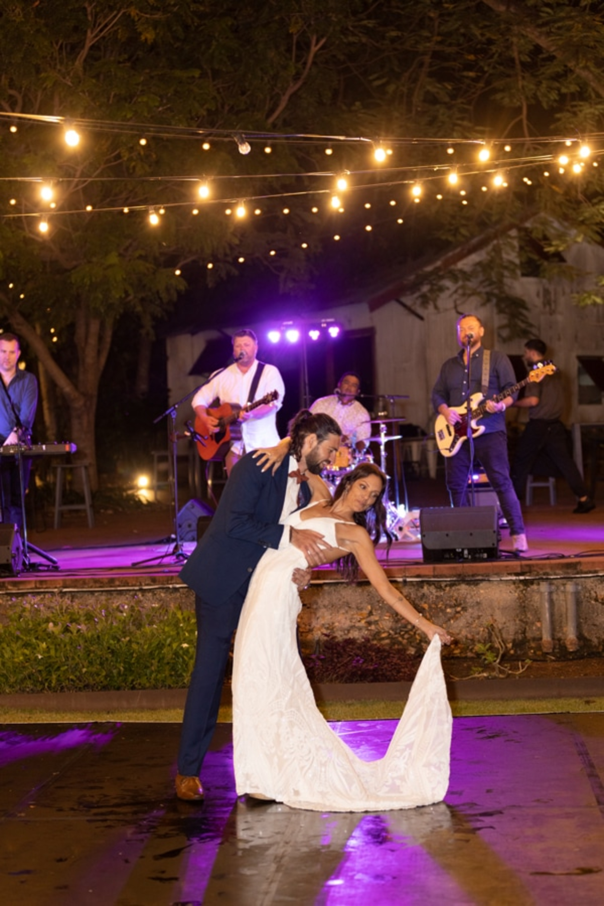 Couple shares a romantic dip on the dance floor as a live band plays under string lights at an outdoor evening wedding.