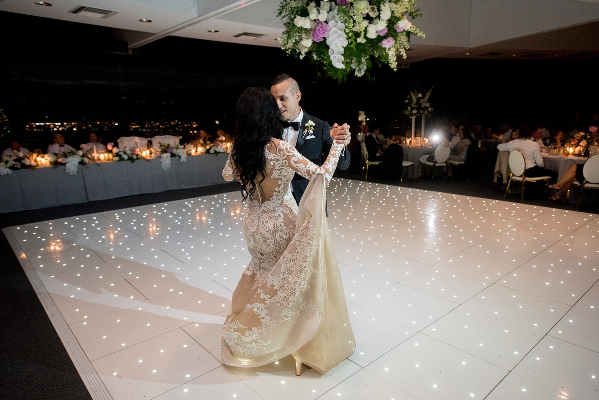 Bride and groom share their first dance on a sparkling white dance floor at an elegant indoor wedding reception.