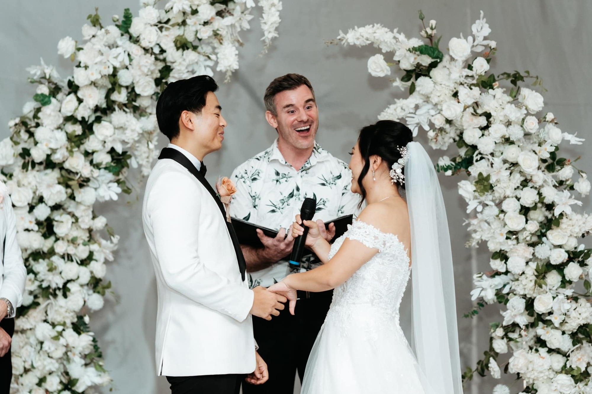 Bride and groom share vows with a smiling officiant beneath a white floral wedding arch.