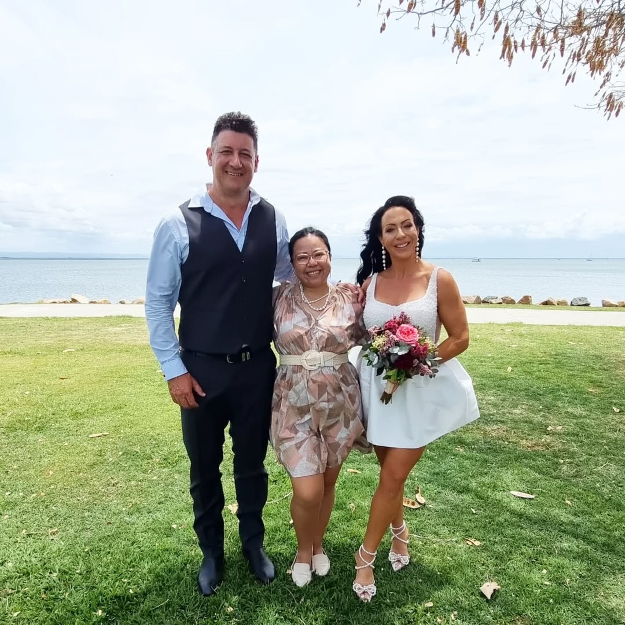 Smiling couple and officiant pose together on a grassy waterfront after a casual outdoor wedding ceremony.