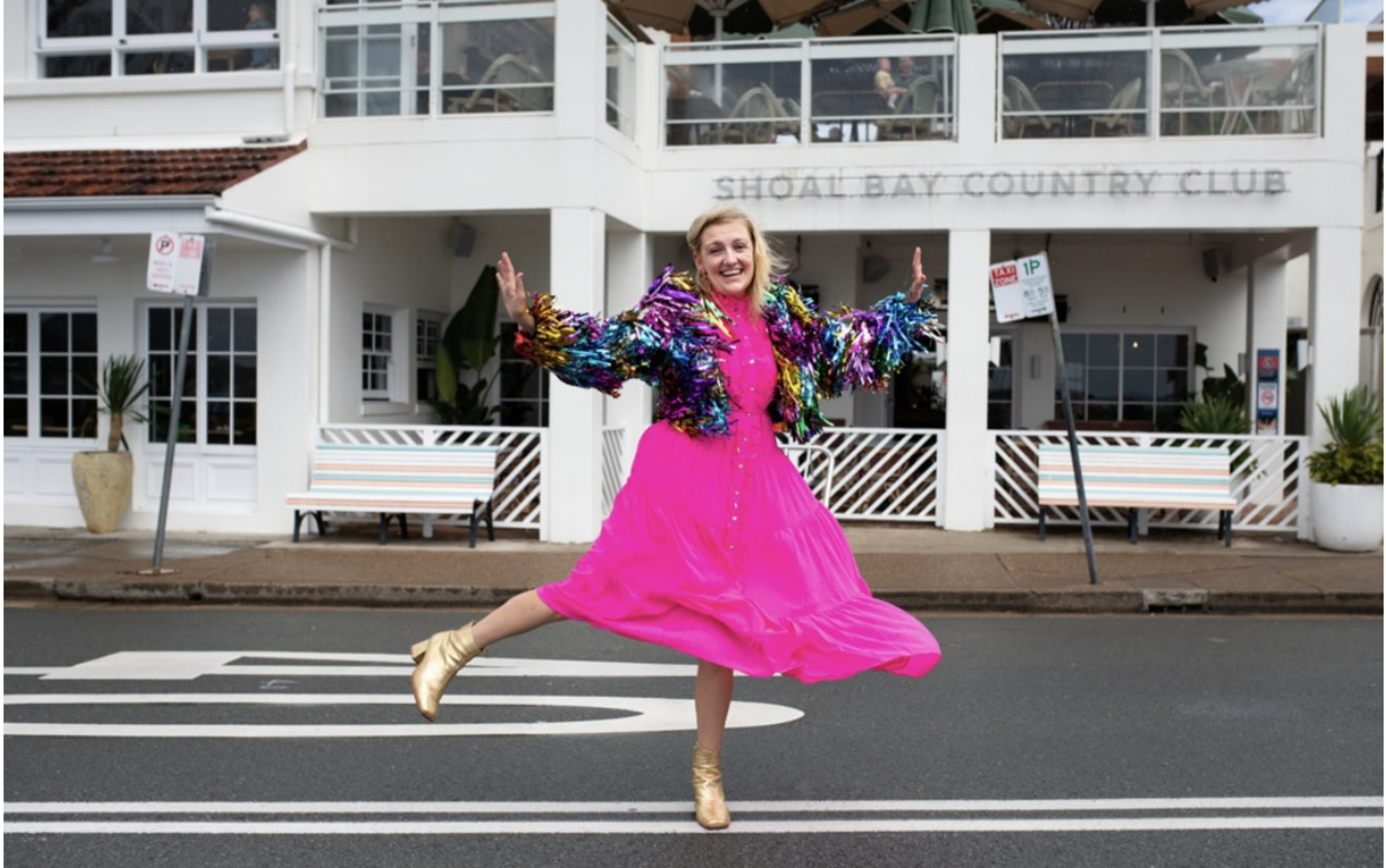 A joyful woman in a bright pink dress and colorful jacket jumps on the street in front of Shoal Bay Country Club.