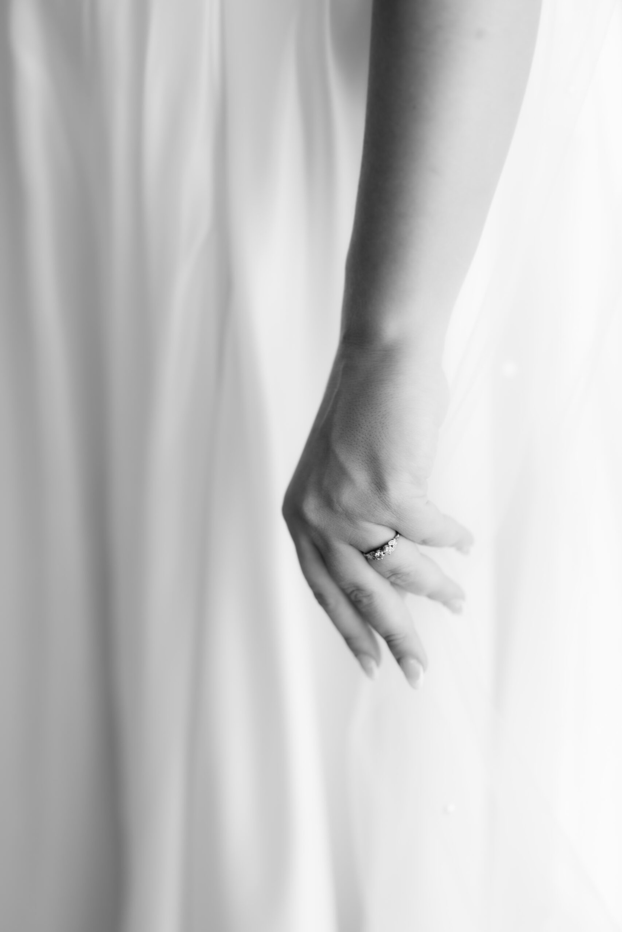 Close-up black and white photo of a bride’s hand resting by her gown, showing a diamond engagement ring.