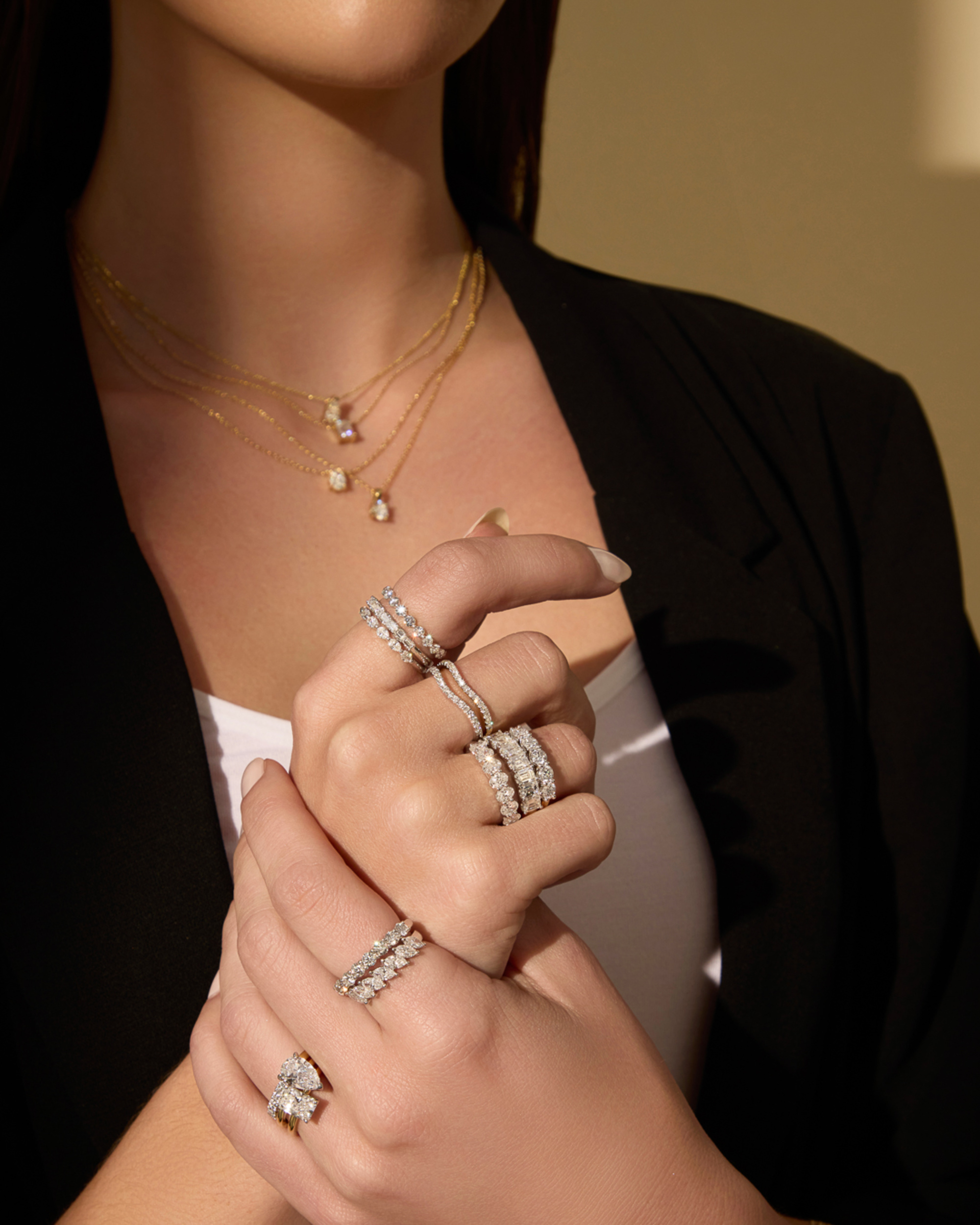 Close-up of a woman wearing stacked diamond rings and layered gold necklaces in warm lighting.