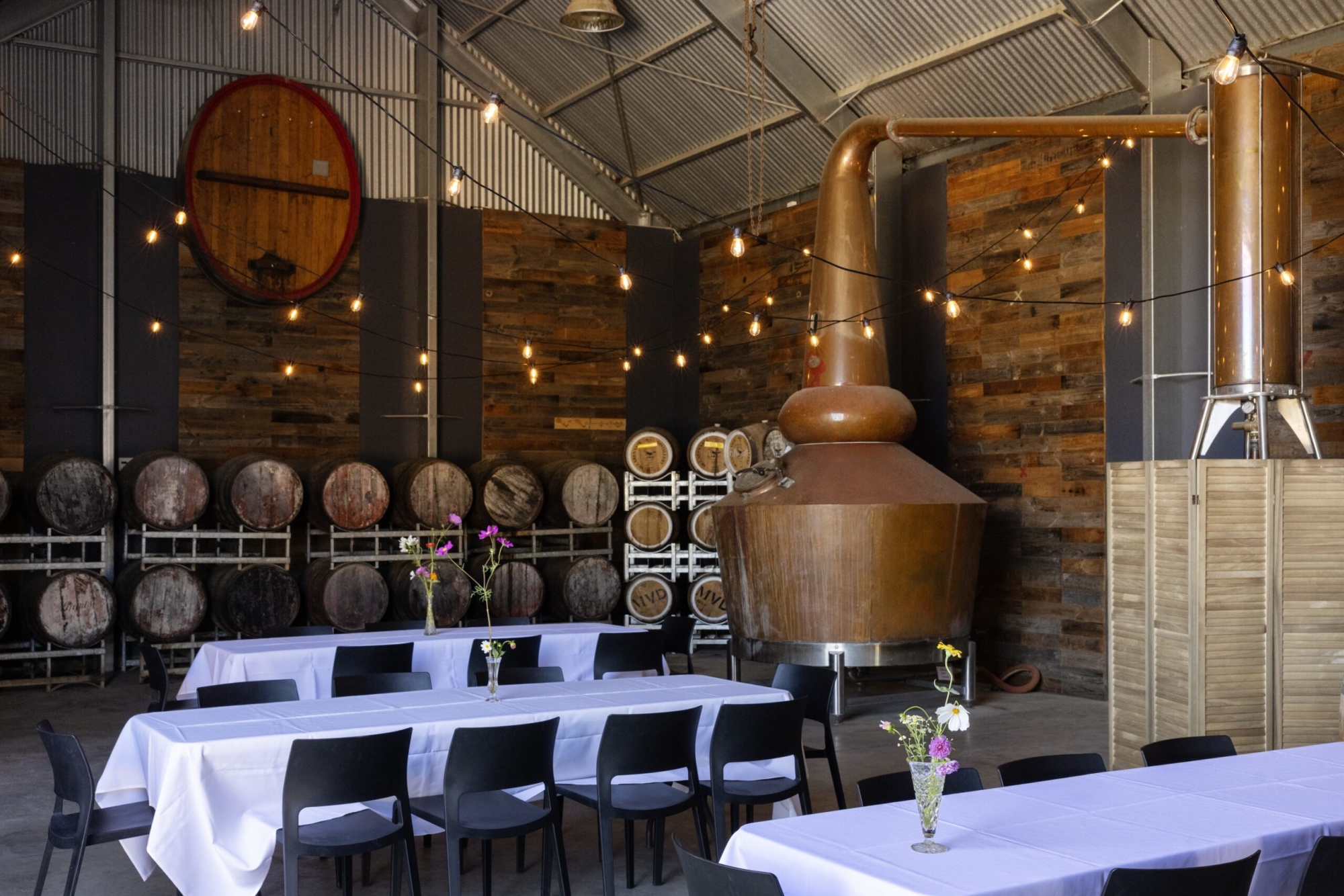 Industrial distillery wedding venue with barrel-lined walls, string lights, and banquet tables with simple floral centerpieces.
